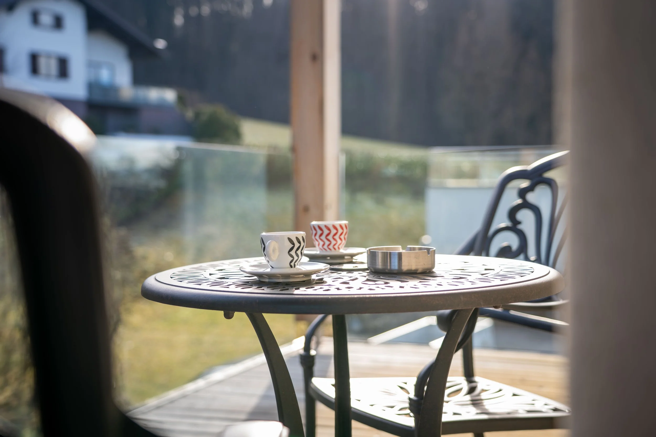 Ein runder Balkon mit einem kleinen Metalltisch, auf dem zwei Kaffeetassen mit Mustern und eine Metalldose stehen, vor einer Glaswand mit Blick auf ein Haus und eine grüne Landschaft bei Sonnenlicht.