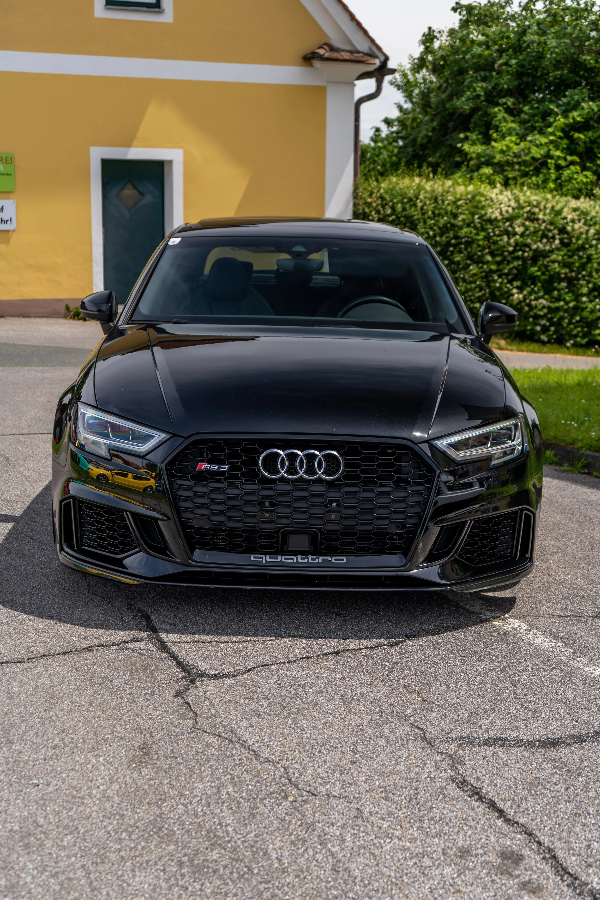 Schwarzer Audi RS 3 Sportback vor einem gelben Haus mit grünem Garten. Auf der Motorhaube ist ein Logo, unter dem eine Leiste mit der Aufschrift ‚quattro‘ ist.