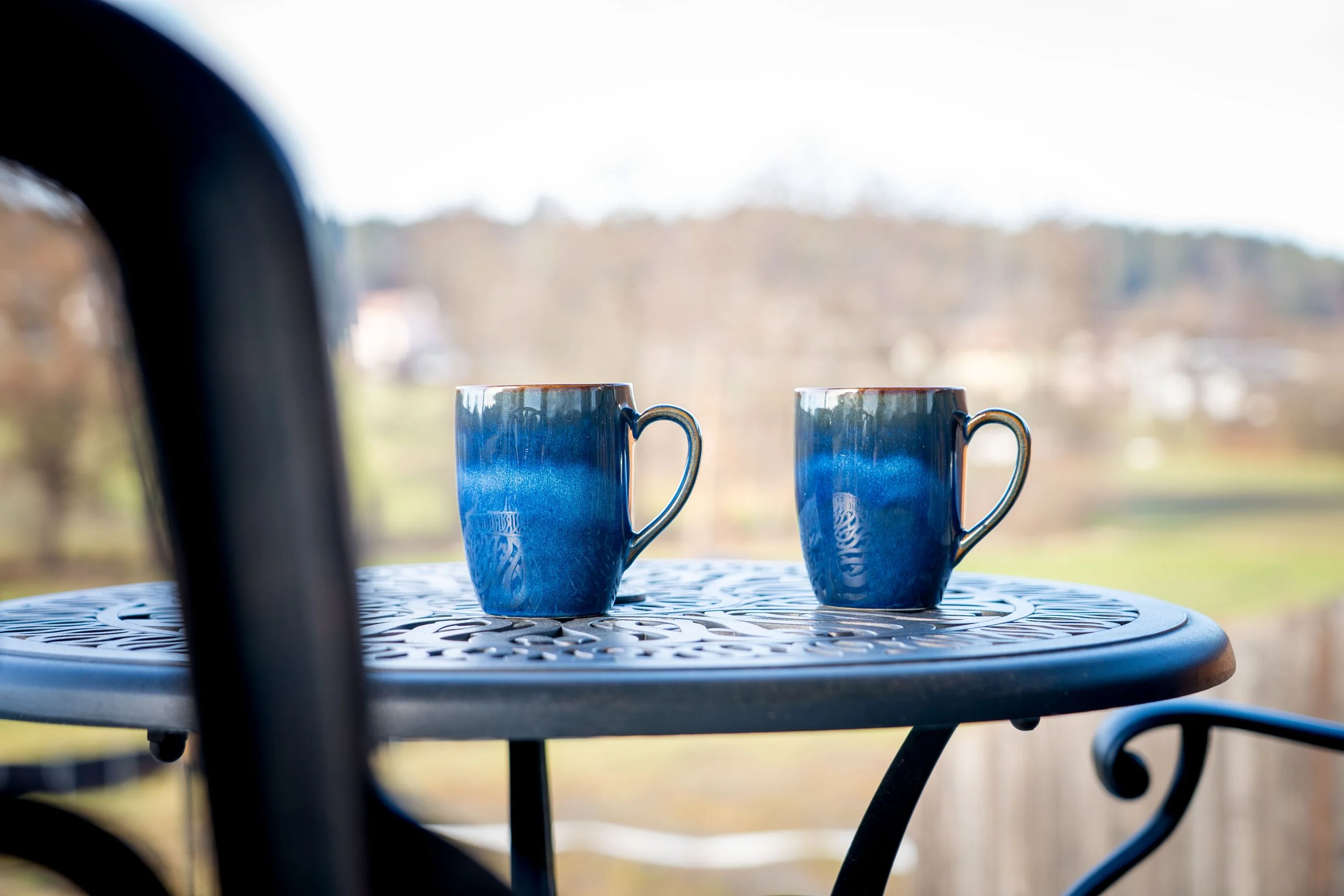 Zwei blaue Kaffeetassen auf einem runden, weißen, gitterartigen Tisch, auf einem Balkon mit Blick auf eine Landschaft aus Bäumen und Hügeln im Hintergrund.
