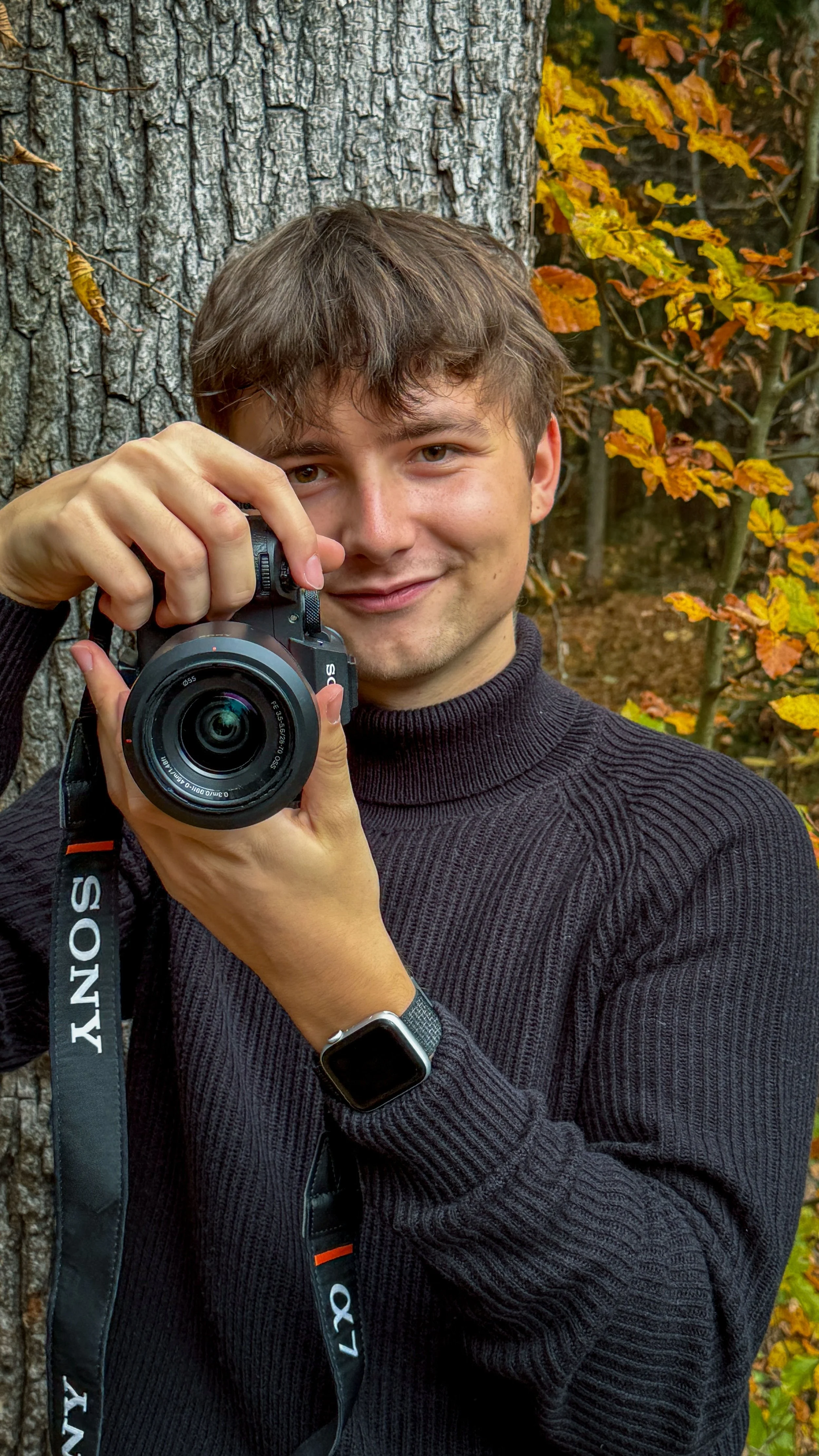 A young man holding a camera, standing outdoors next to a tree and colorful autumn leaves.