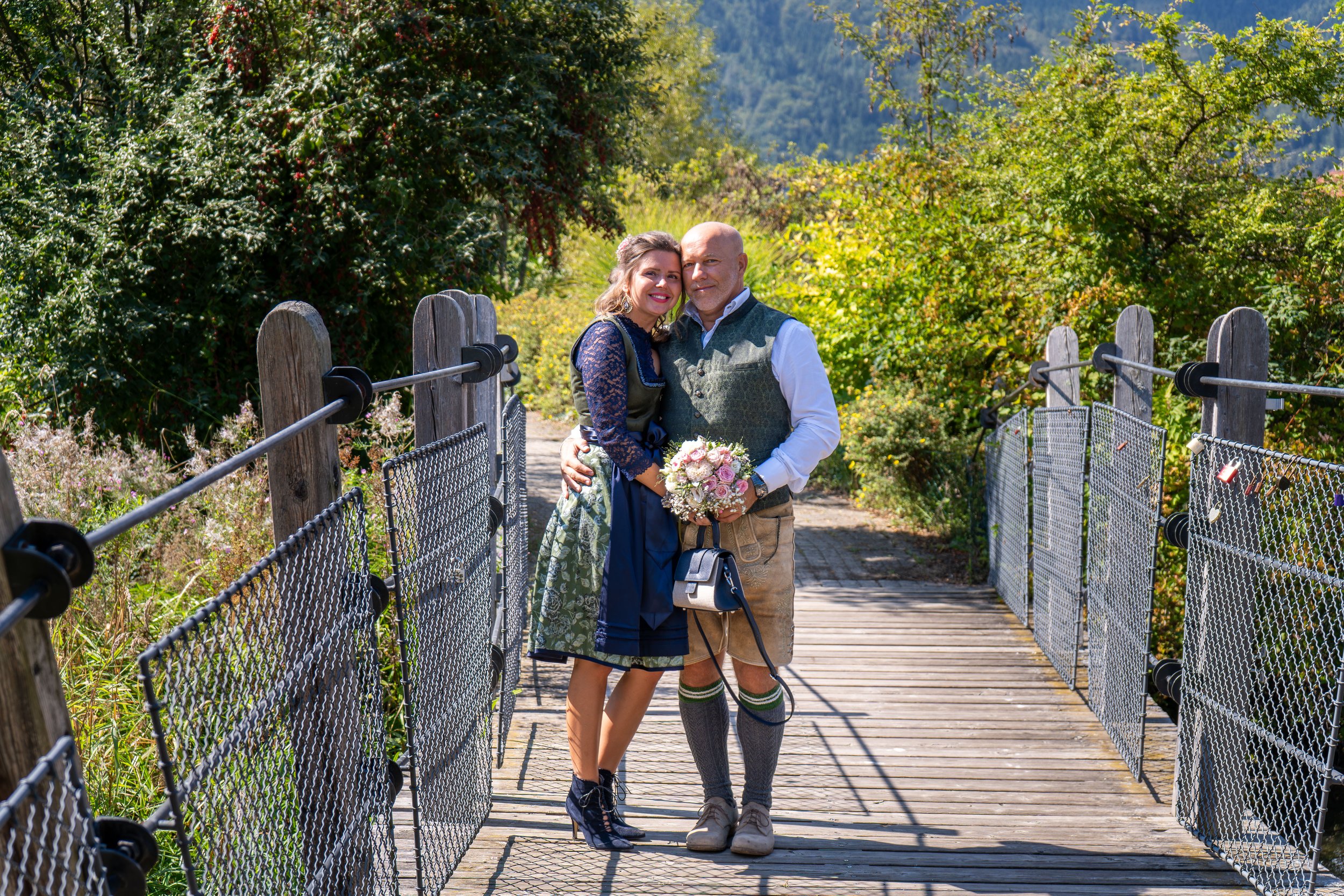 Ein Paar in traditionellen bayerischen Trachten steht auf einer Brücke in einer Naturlandschaft mit Bäumen und Bergen im Hintergrund, während sie sich umarmen und einander zu lächeln. Die Frau hält einen Blumenstrauß.