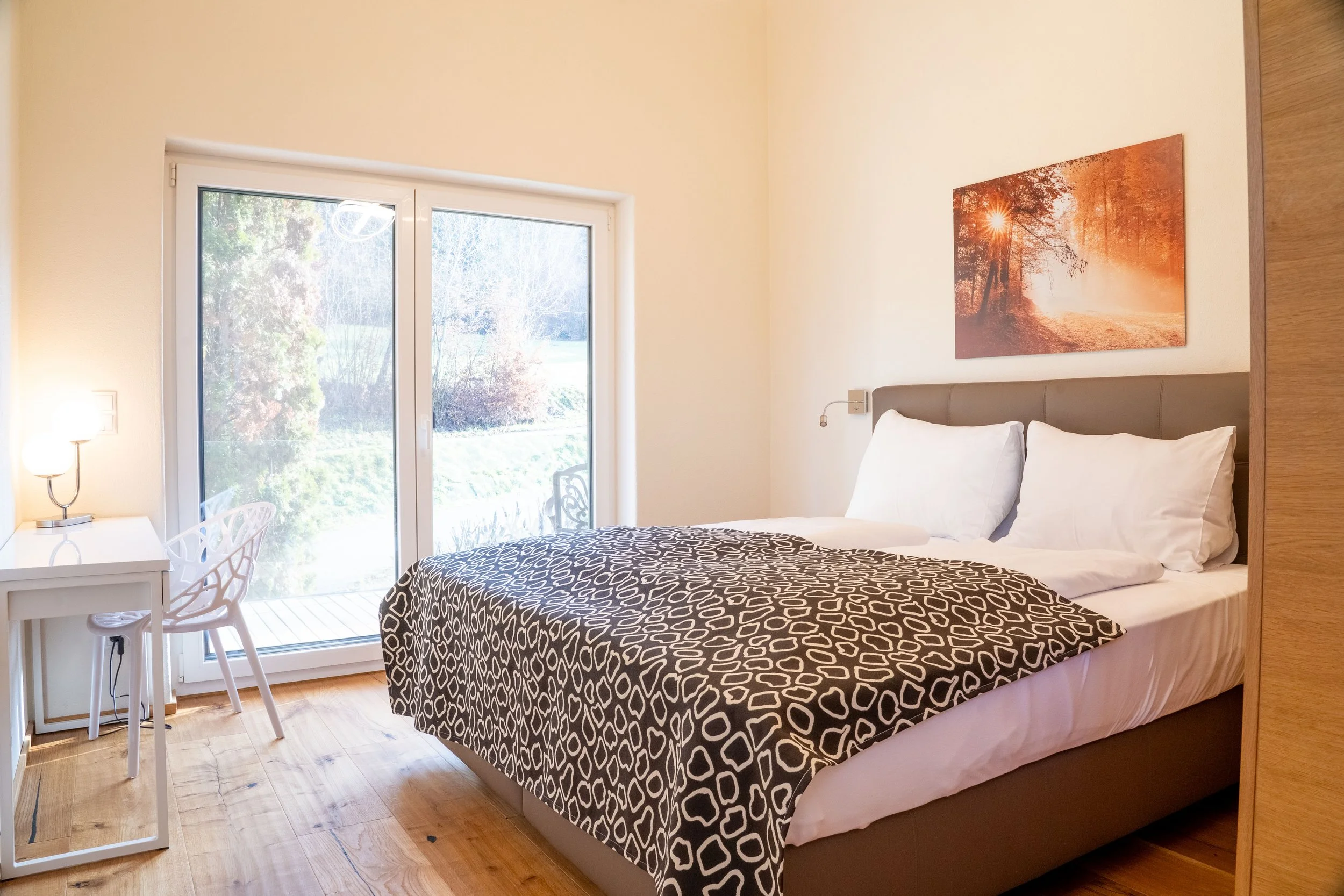 Ein Schlafzimmer mit einem Bett, weißen Bettlaken, schwarzen und weißen Bettbezug, einem Nachttisch mit einer Lampe, einem Schreibtisch mit einem Stuhl und einem großen Fenster mit Blick nach draußen auf einen Garten.