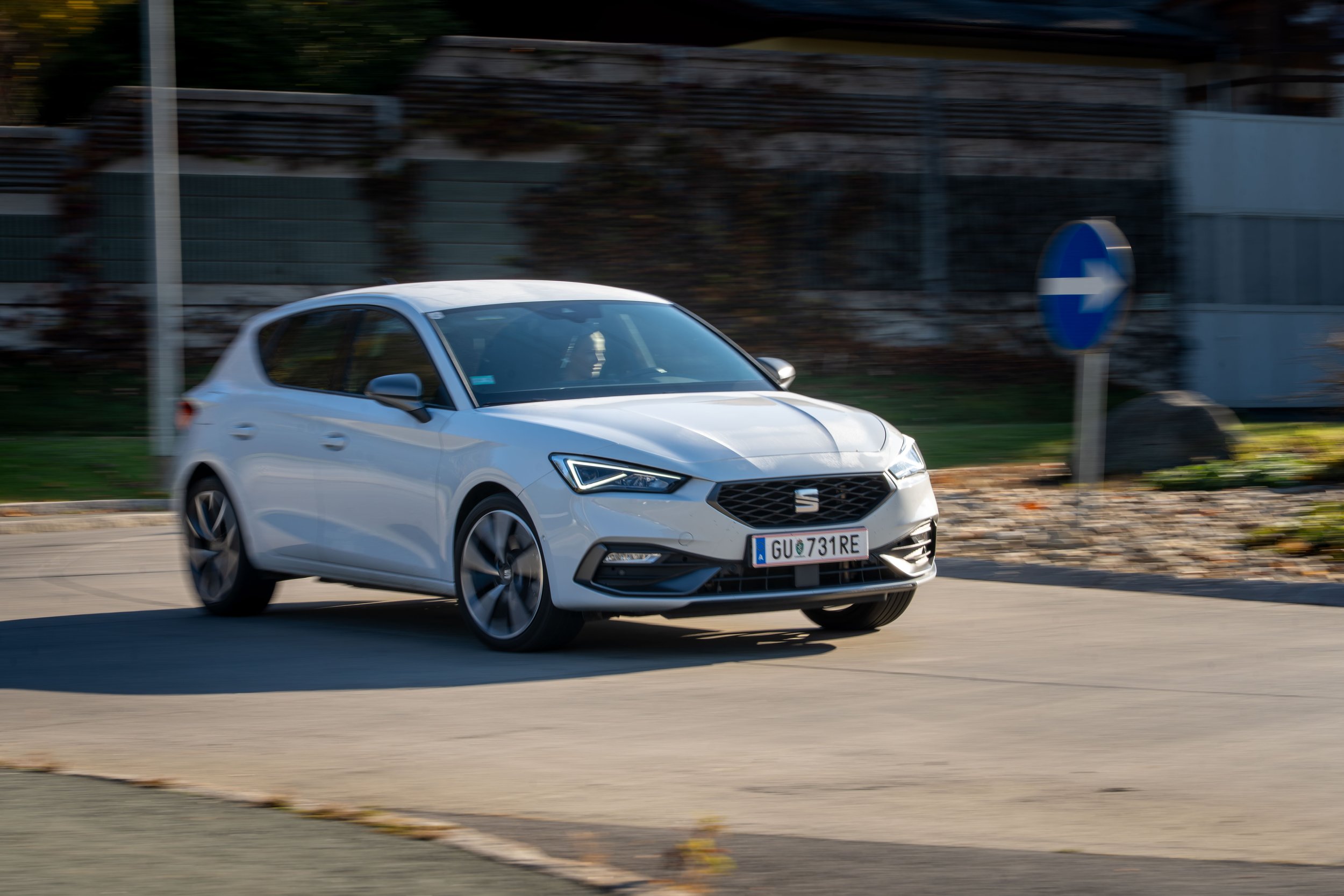 White modern hatchback car driving on a road near a blue right-turn traffic sign, with fence and foliage in the background.