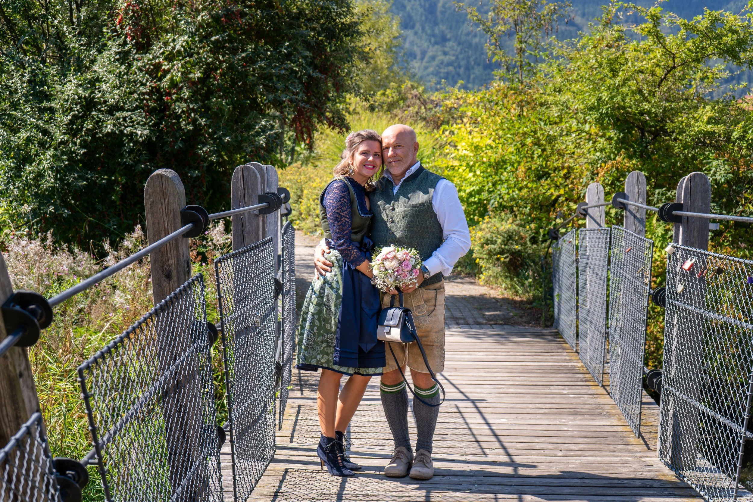A couple dressed in traditional German attire standing on a wooden bridge, smiling, with a lush green landscape in the background.