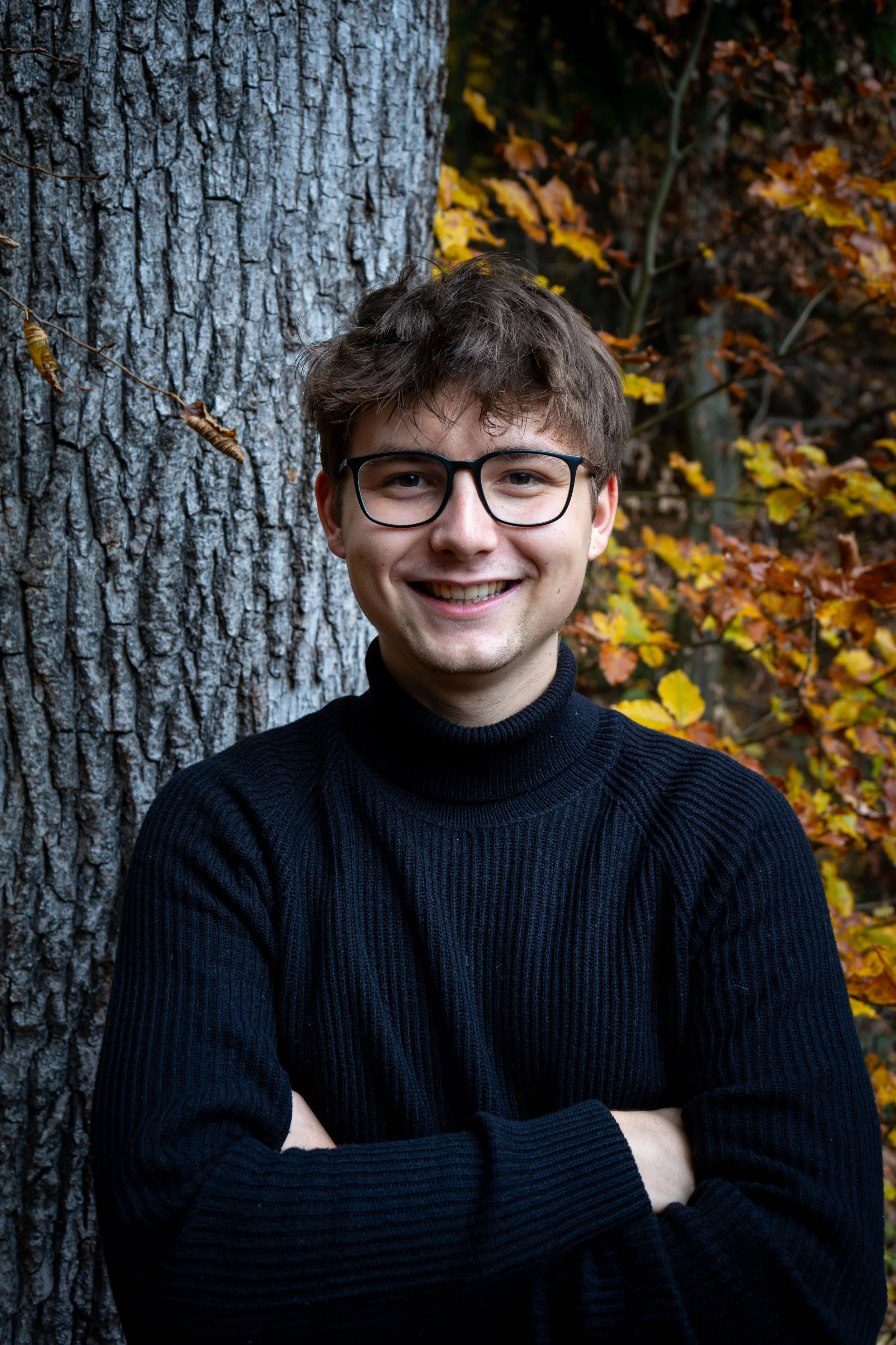 A smiling young man with glasses and brown, tousled hair standing outdoors in front of a large tree trunk and autumn-colored leaves. Ein lächelnder junger Mann mit Brille und braunem, zerzaustem Haar steht im Freien vor einem großen Baumstamm in Graz