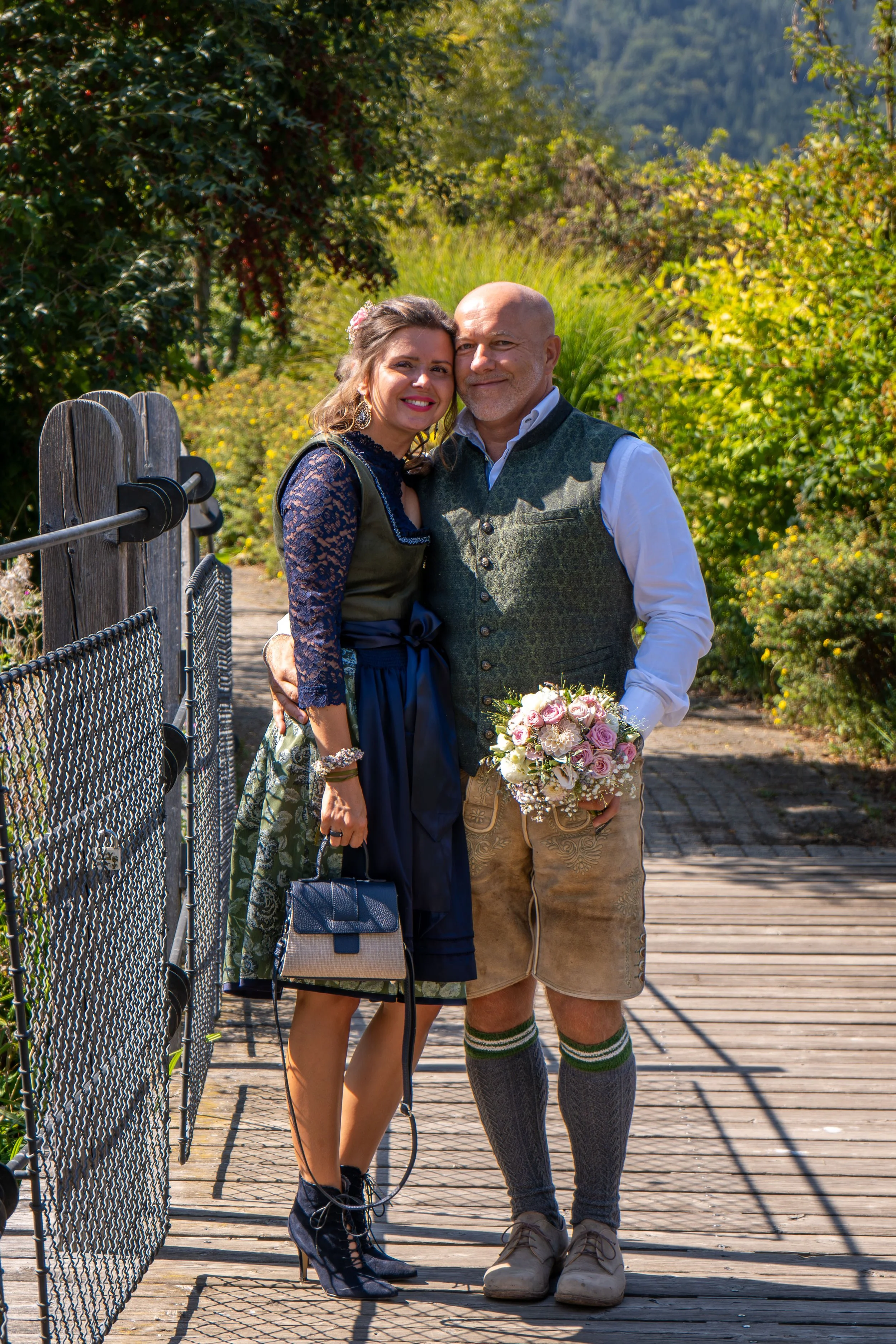 A man and woman standing close together on a wooden bridge, smiling and holding a bouquet of pink and white roses, surrounded by lush green trees and hills in the background.