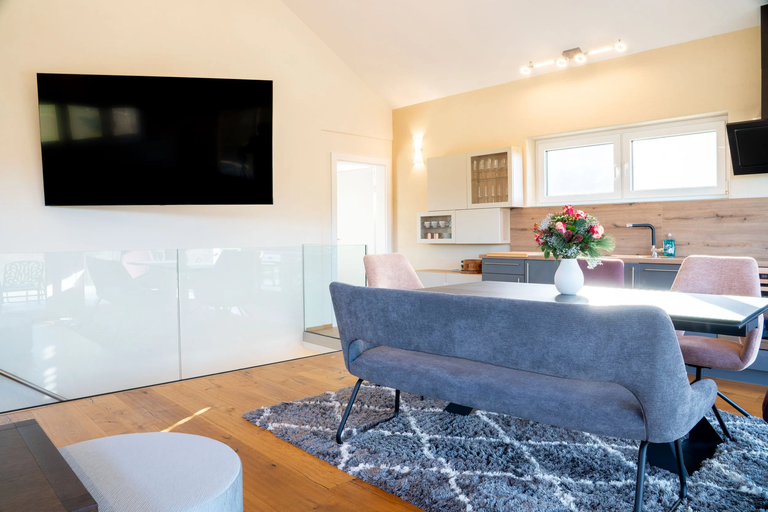 Modernes Wohnzimmer mit grauem Sessel, hellem Holzfußboden, Teppich, KV-Küche mit weißen Schränken, Blumenstrauß auf Tisch, große Fenster, Wandlampen, Fernseher