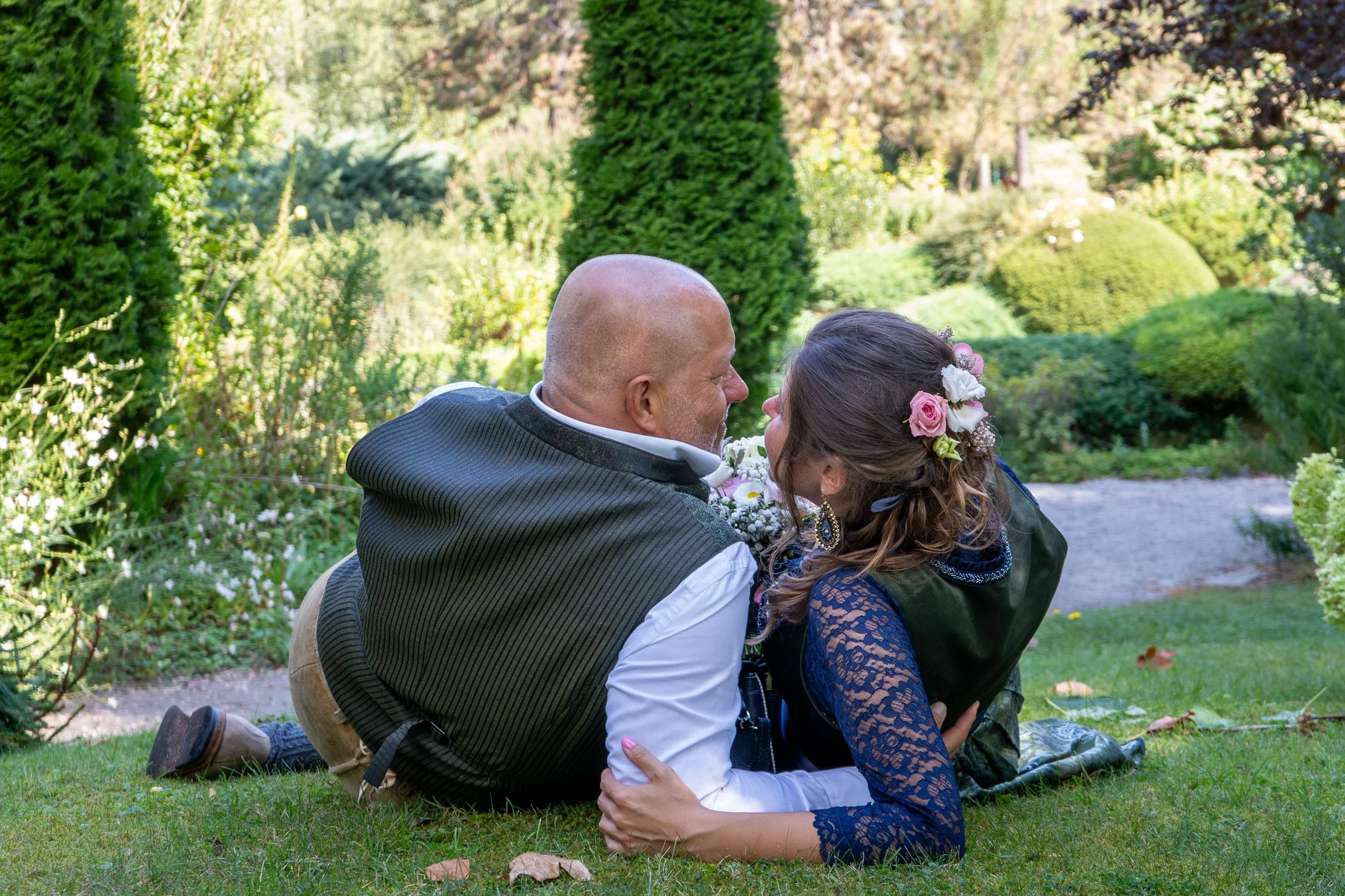 A man and woman sitting on the grass in a garden, kissing. The woman has flowers in her hair, and the man is bald. They are surrounded by lush greenery and trees.