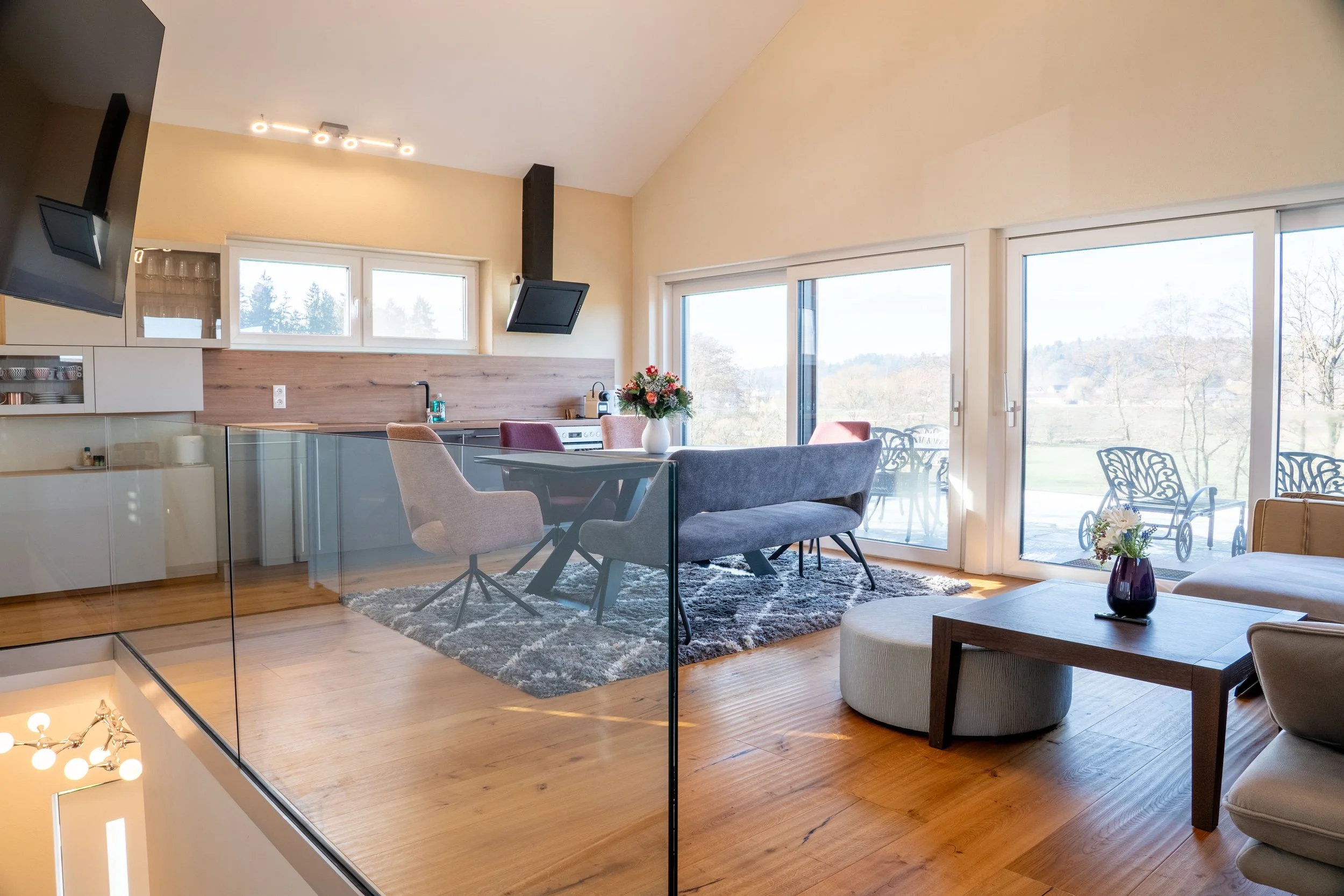 A modern living room with large sliding glass doors showing a scenic outdoor view. The room has a high ceiling with a light fixture, a wooden accent wall with a kitchen sink, a dining table with a flower vase, and a cozy sitting area with a sofa, arm