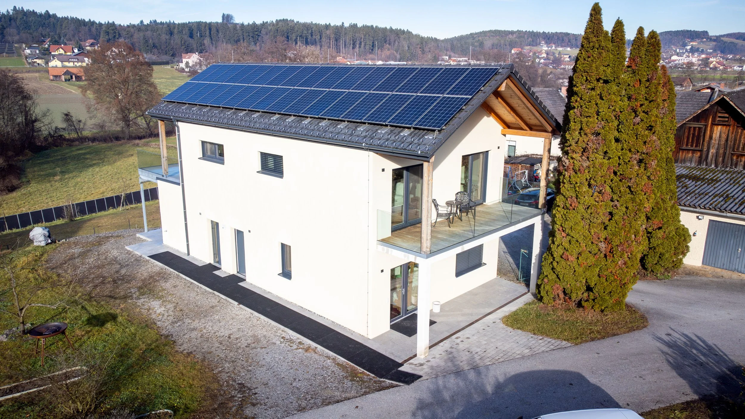 Ein modernes Haus mit Solarpanelen auf dem Dach, einem Balkon mit Möbeln, umgeben von Bäumen und in einer ländlichen Gegend.