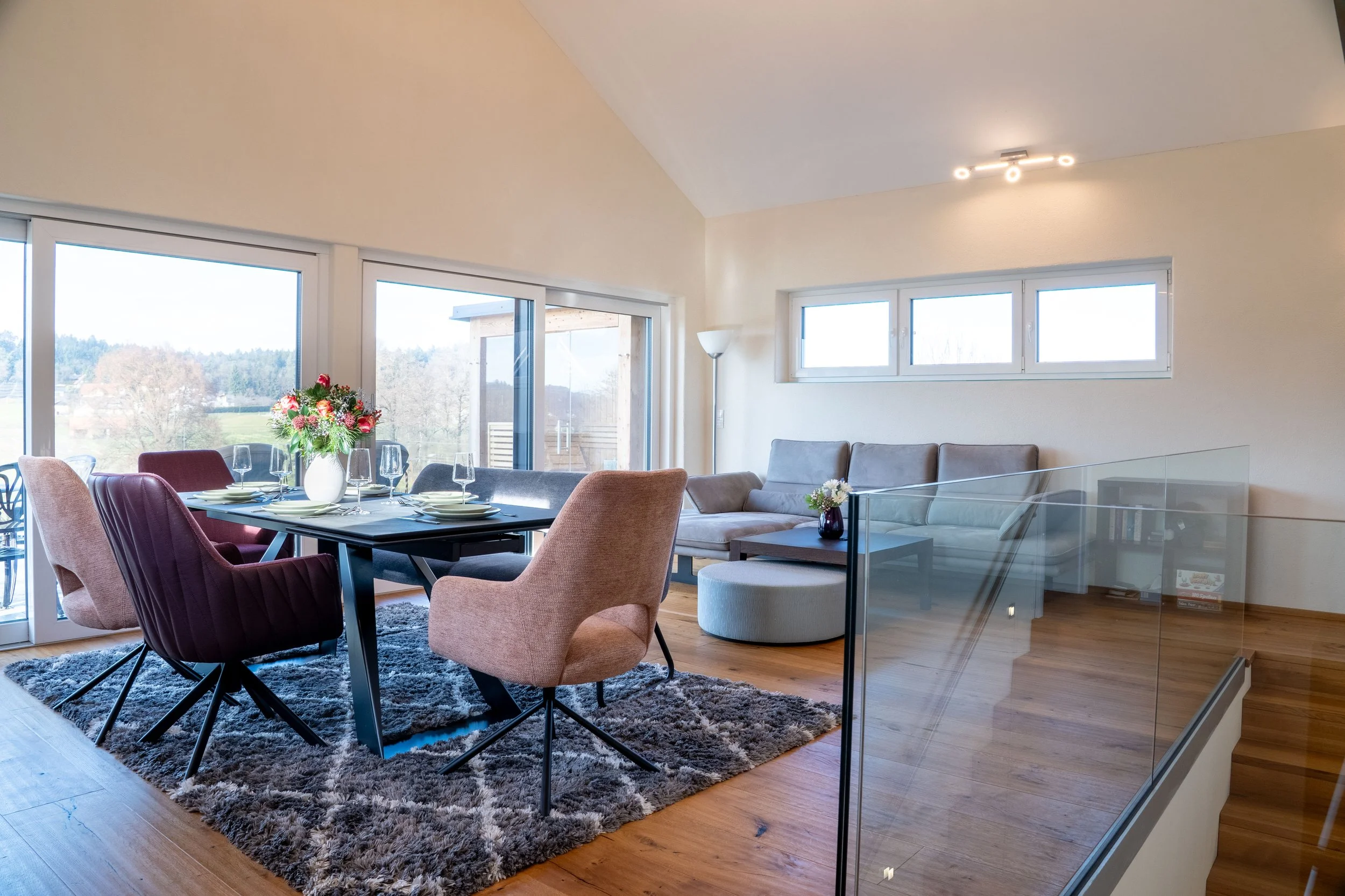 A bright, modern living and dining area with large windows showcasing a scenic outdoor view. The dining table is set with plates, wine glasses, and a floral centerpiece. There is a gray sofa, a small side table with a flower vase, a tall floor lamp, 