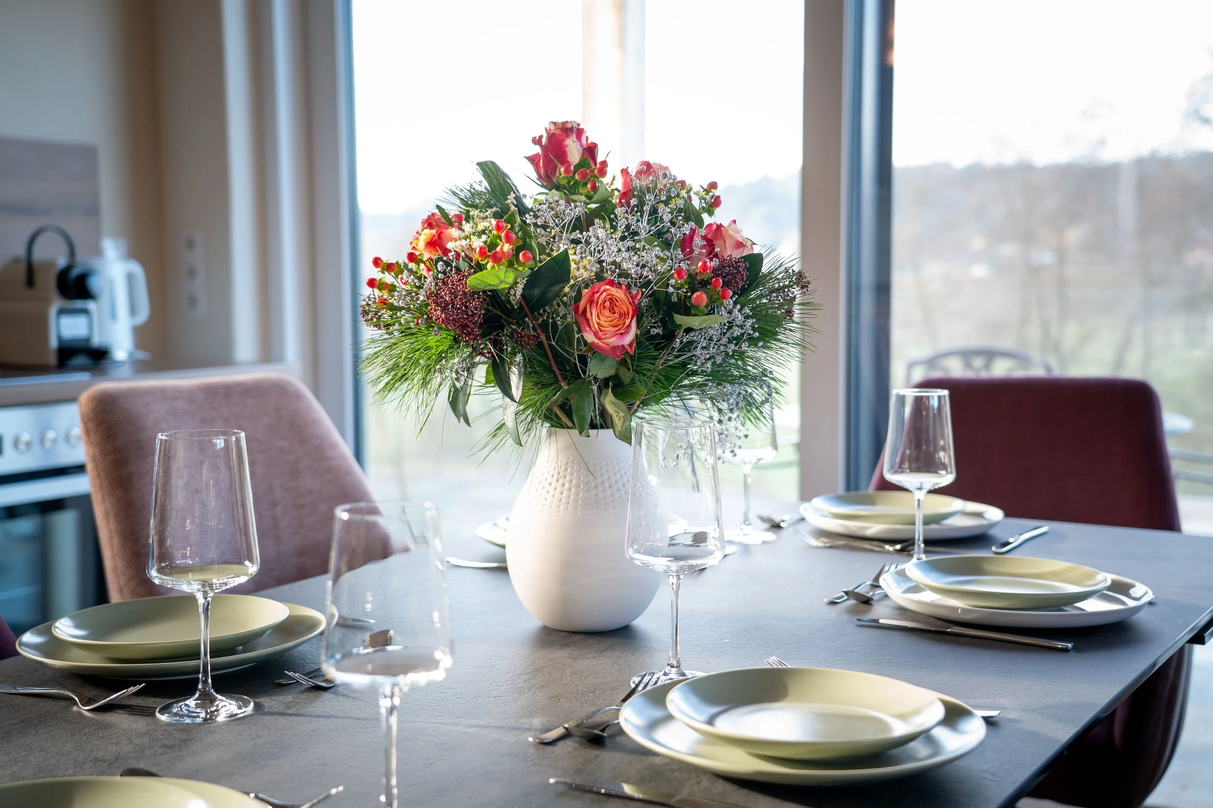 A dining table with a floral centerpiece, wine glasses, and tableware in a room with large windows.