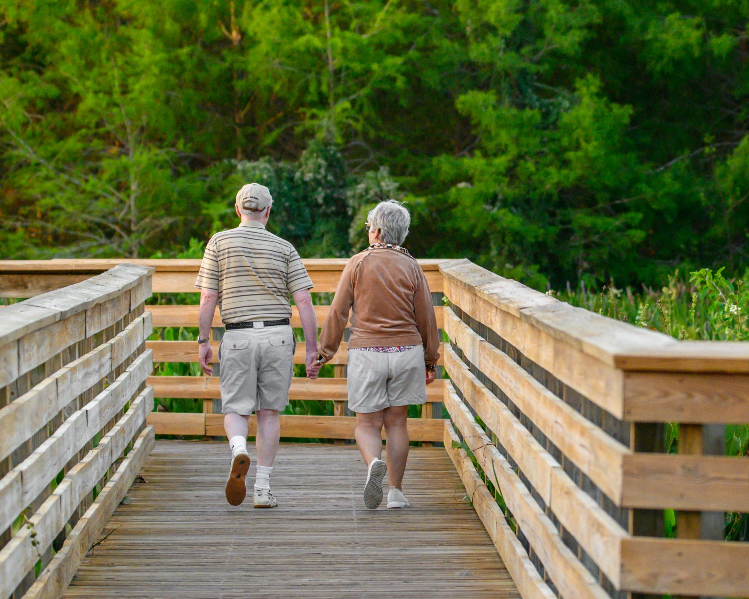 An elderly couple walking hand in hand on a wooden bridge in a green, leafy outdoor setting.