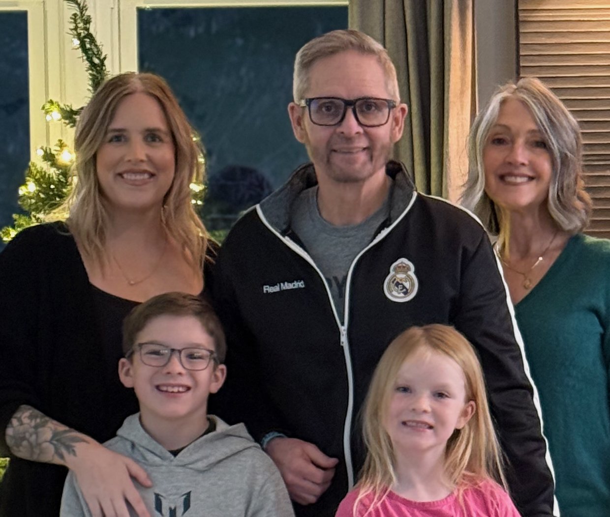 Group of five people, two children and three adults, smiling indoors near a Christmas tree, holiday decorations, and wooden window blinds.