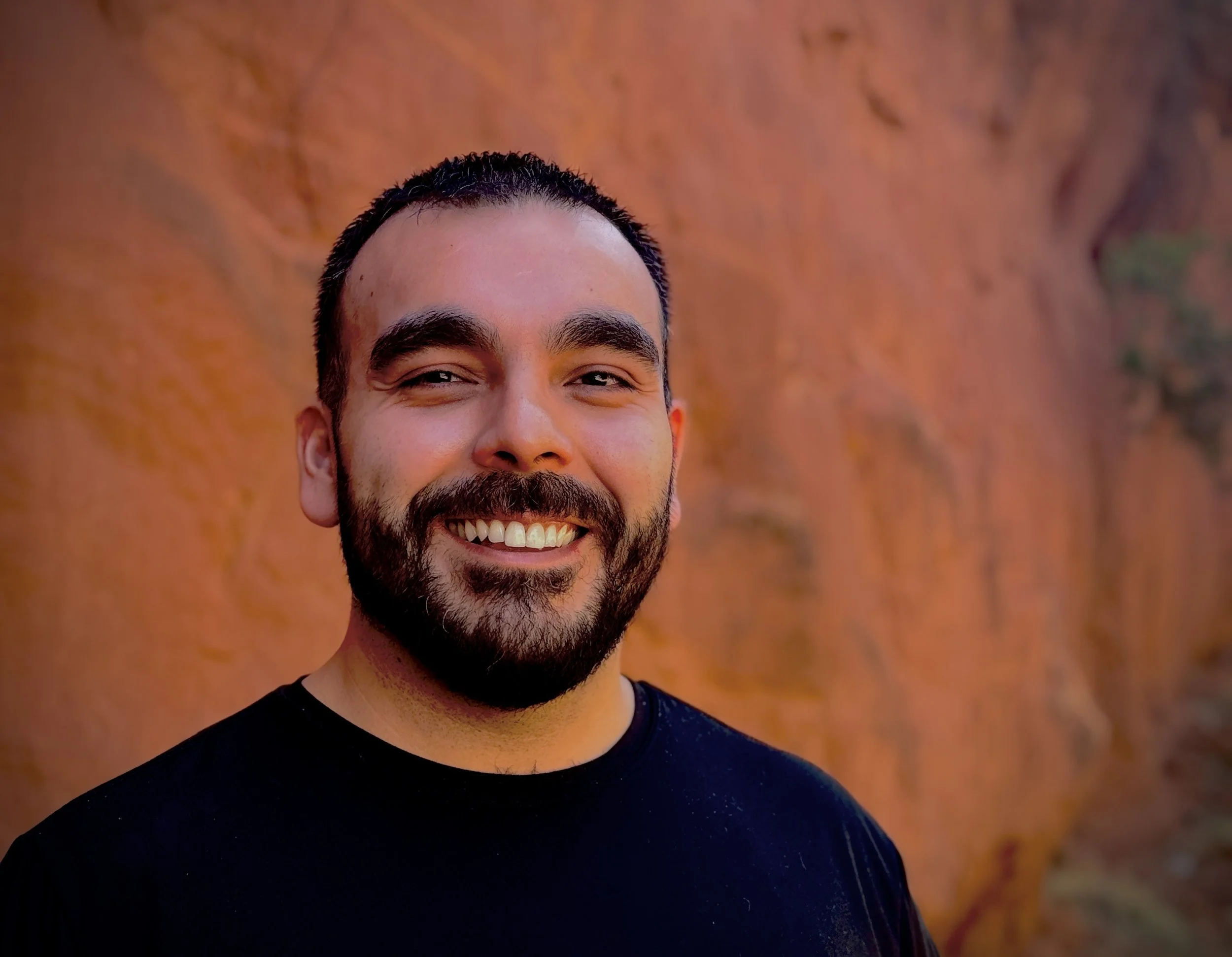 Close-up of a smiling man with dark hair, beard, wearing a black shirt, standing against a brownish-red textured wall.