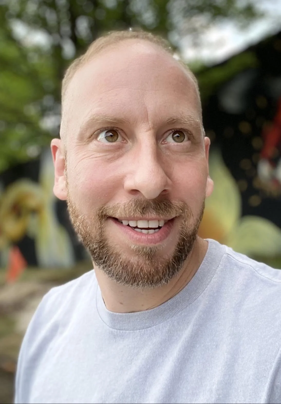 A close-up of a smiling man with a beard and short hair, outdoors with trees and colorful blurred background.