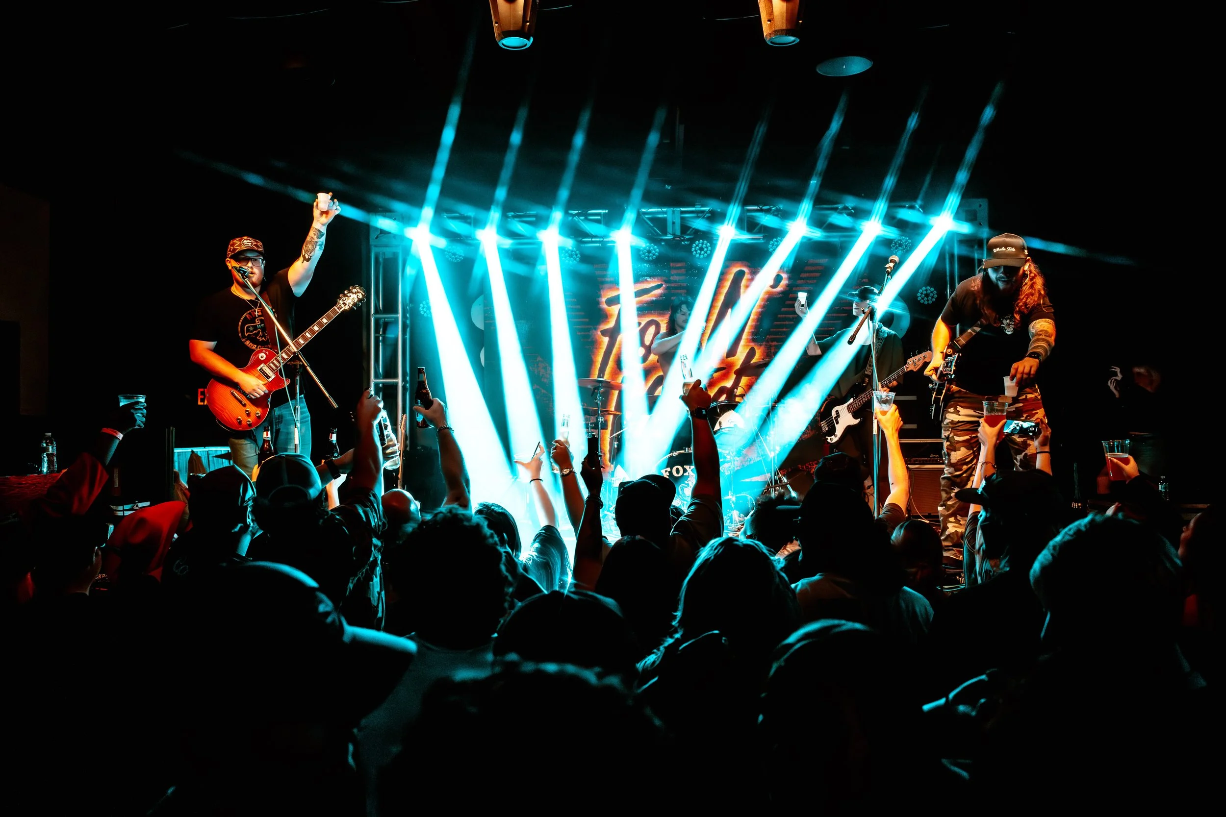 Three men shopping at a merchandise stall with clothing and vinyl records, inside a dimly lit venue with a sign thanking supporters of live music.