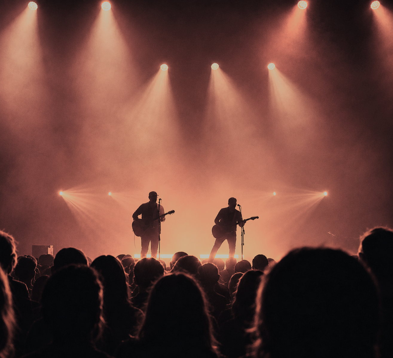 Two musicians with guitars performing on stage under warm orange and pink lights, silhouetted against the bright background, with an audience watching in the foreground.