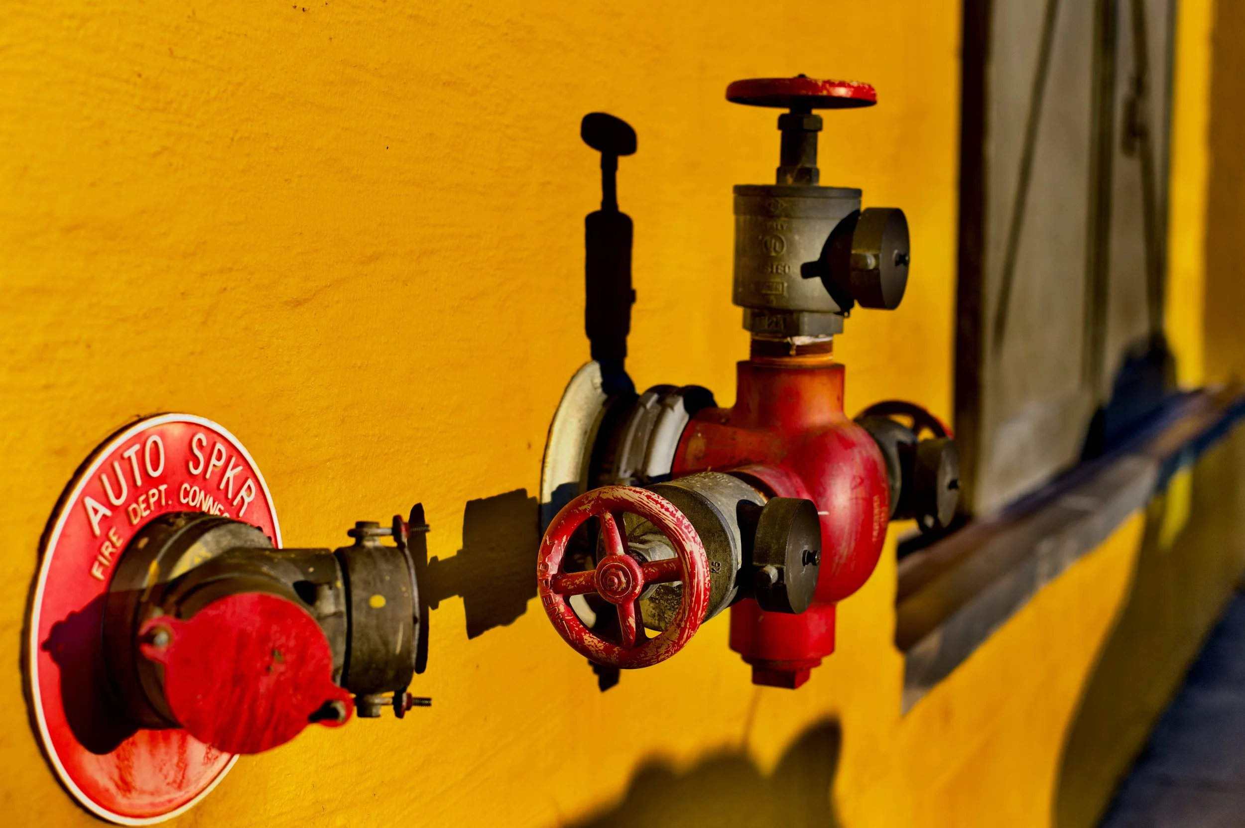 Close-up of red fire department valves and pipes mounted on a yellow wall, with a partially visible red and white circular sticker on the left.