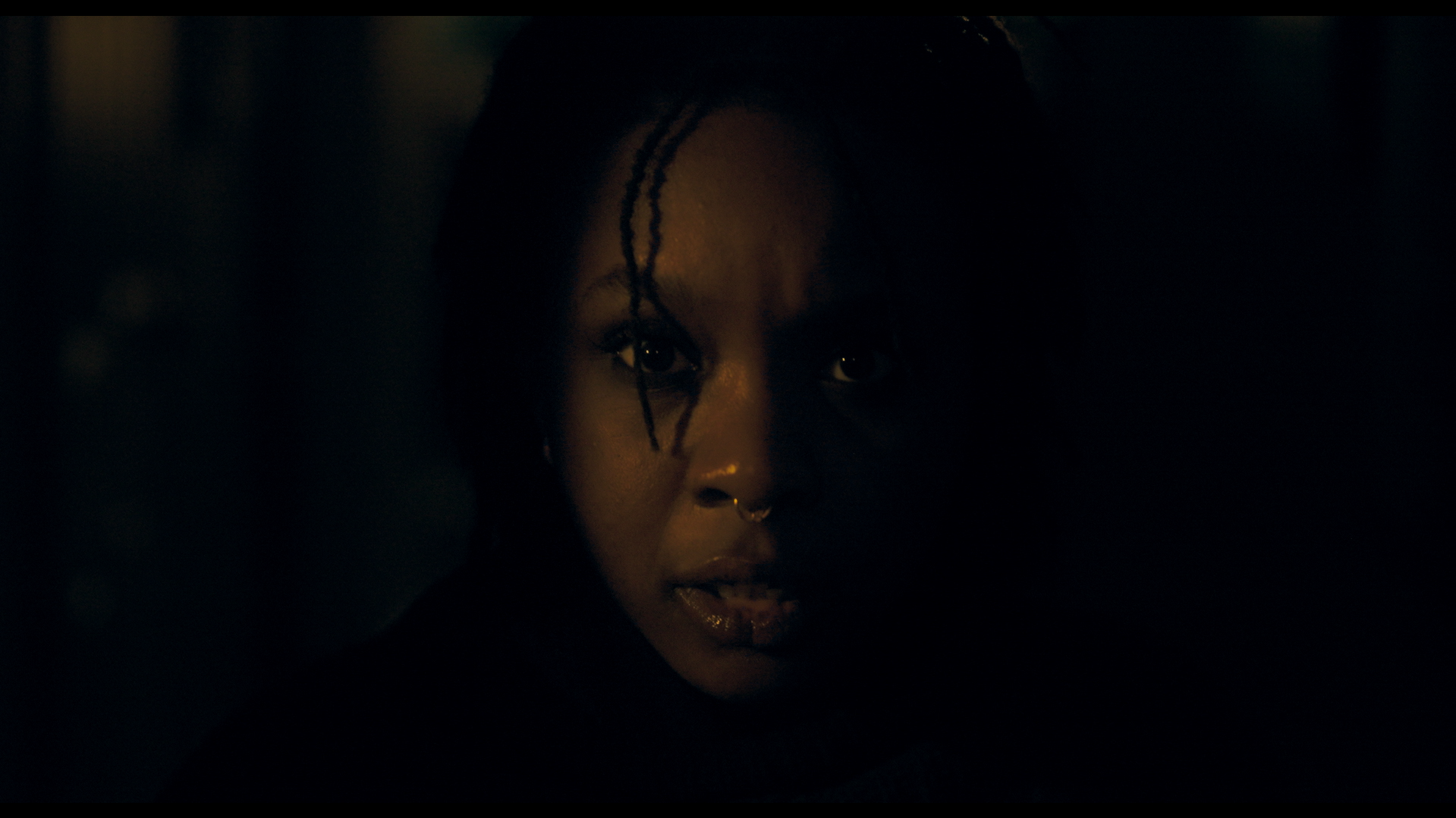 Close-up of a woman's face in darkness, with only part of her face illuminated, showing her eyes, nose piercing, and locs.