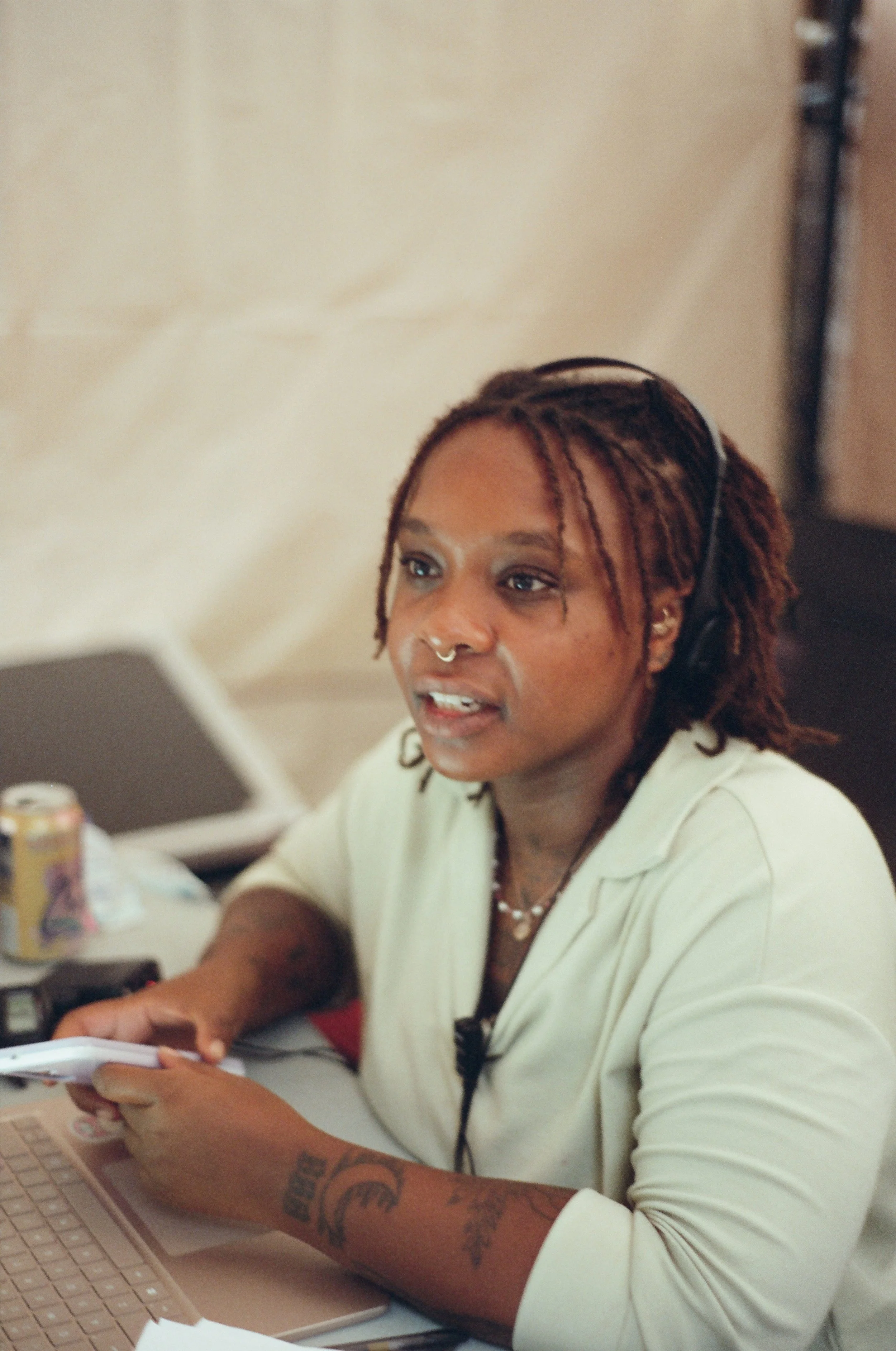 A woman with reddish-brown locs and a septum piercing is sitting at a desk, looking at a computer screen. She is wearing a cream-colored shirt and has visible tattoos on her arm. There are various objects on the desk, including a keyboard, papers, a soda can, and a phone.