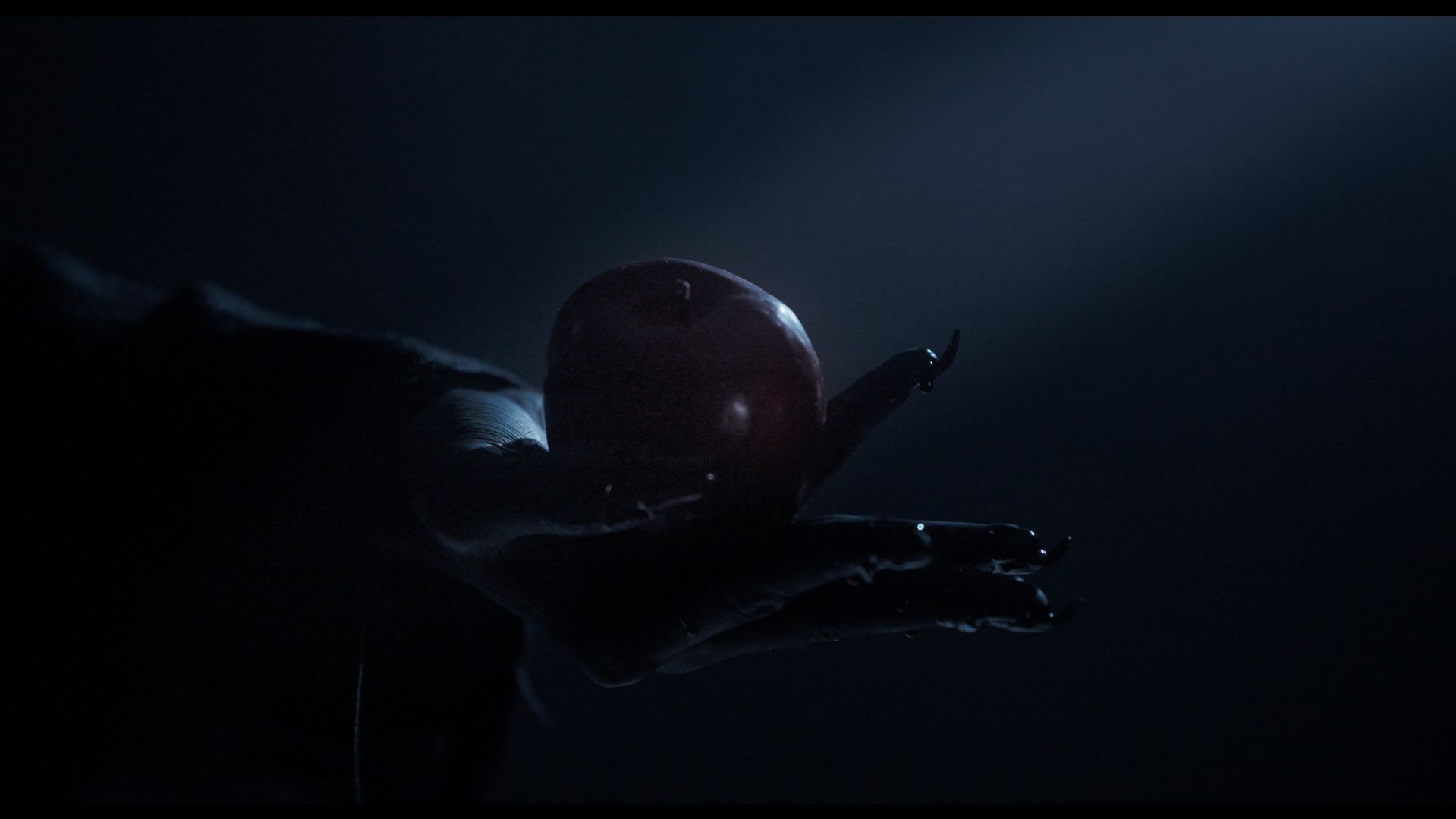 A dark, moody photograph of a hand holding a red apple, with dramatic lighting creating shadows and highlights on the apple and hand.
