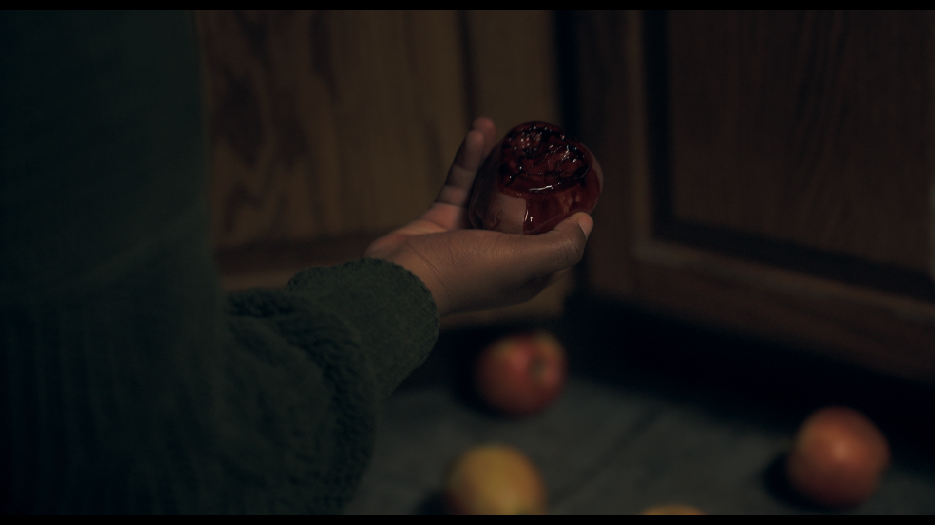 A person holding a rotten, bleeding apple in a dimly lit room. There are apples on the floor underneath and in the background.
