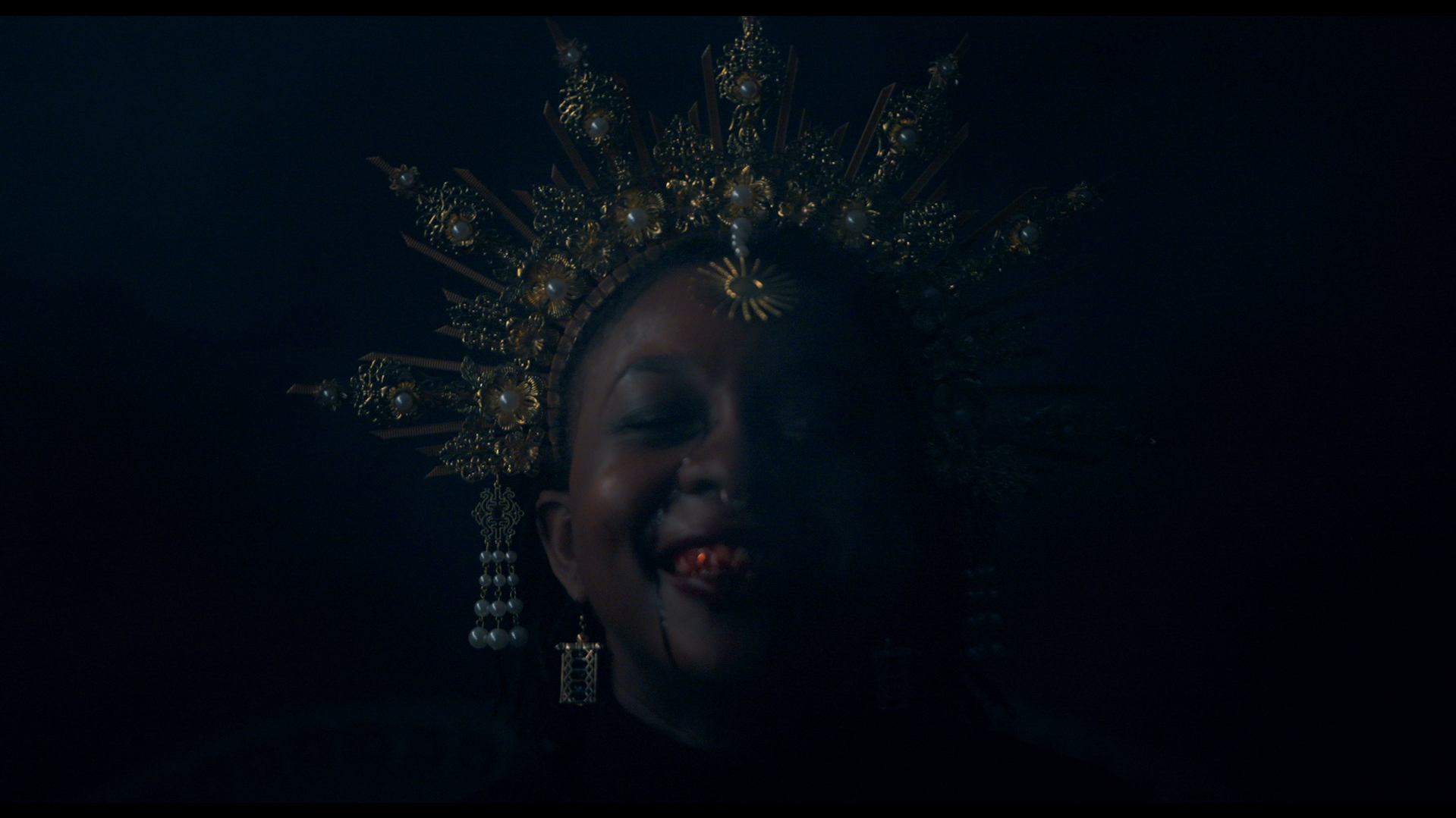 Close-up of a smiling woman with dark skin, wearing an ornate gold crown and earrings, in a dark setting.