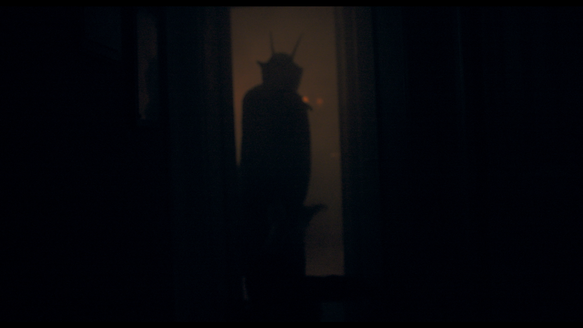 Dark hallway with a mysterious shadowy figure of a person wearing a costume with horns, standing in the doorway, illuminated from behind.