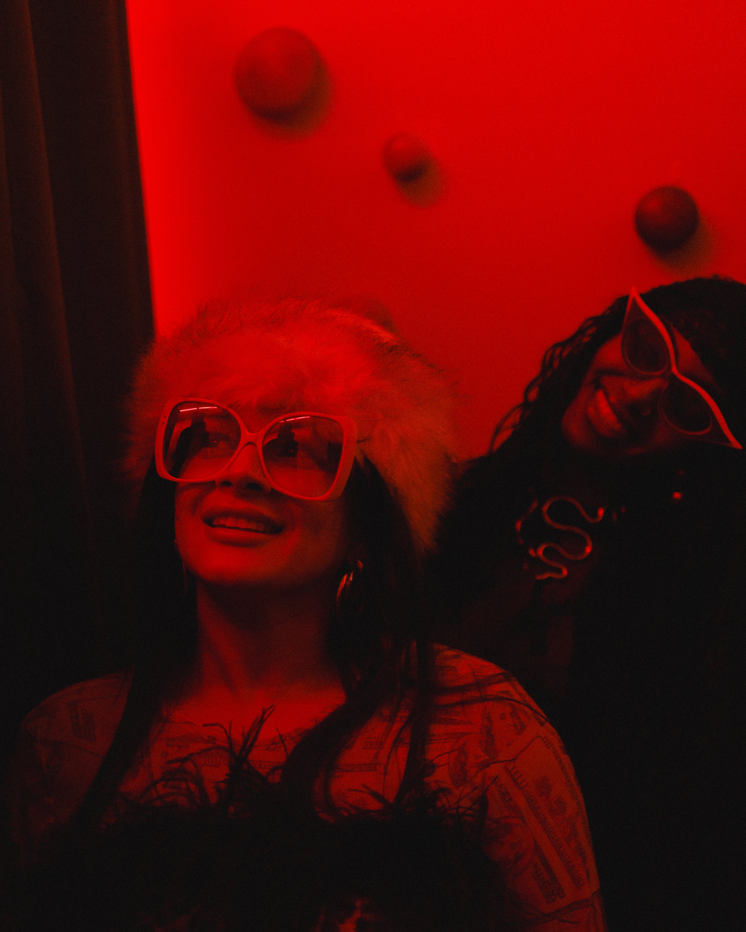 A person smiling wearing sunglasses with a pink frame in a dark environment with red lighting. The person has long dark hair and is wearing a shirt with text and a map pattern.