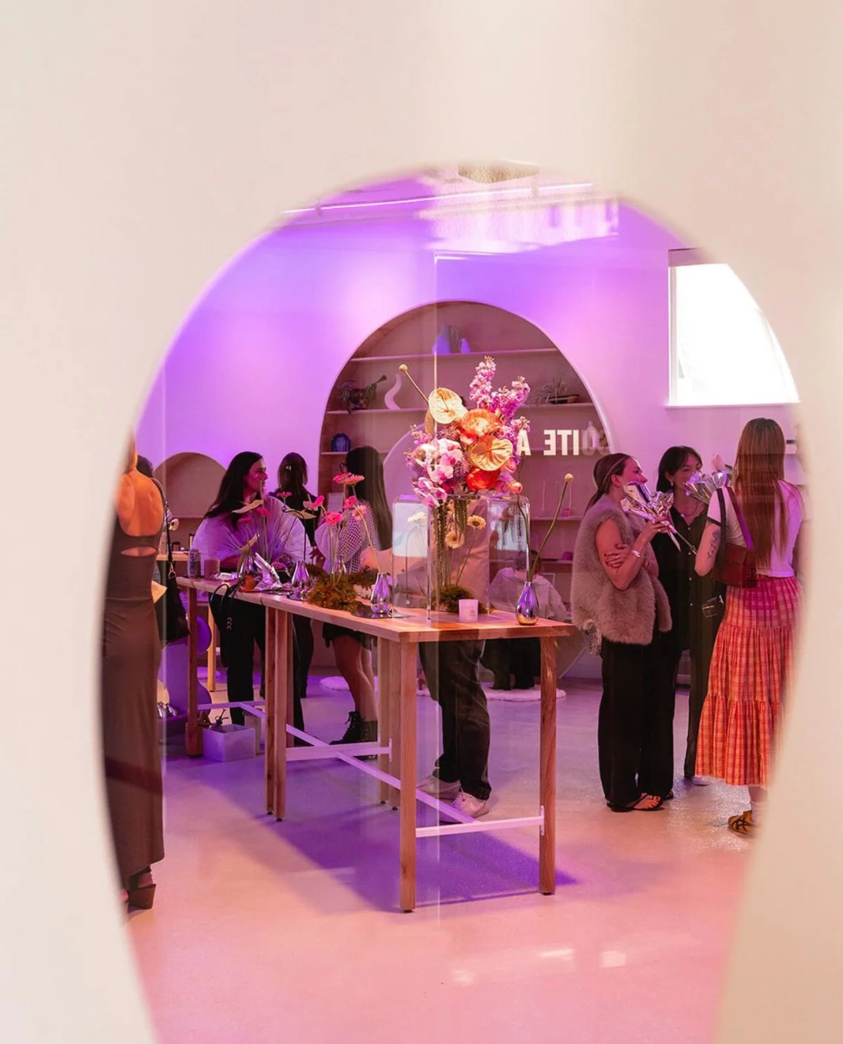 People socializing at a modern, decorated interior space with pink lighting, floral arrangements, and a wooden table.