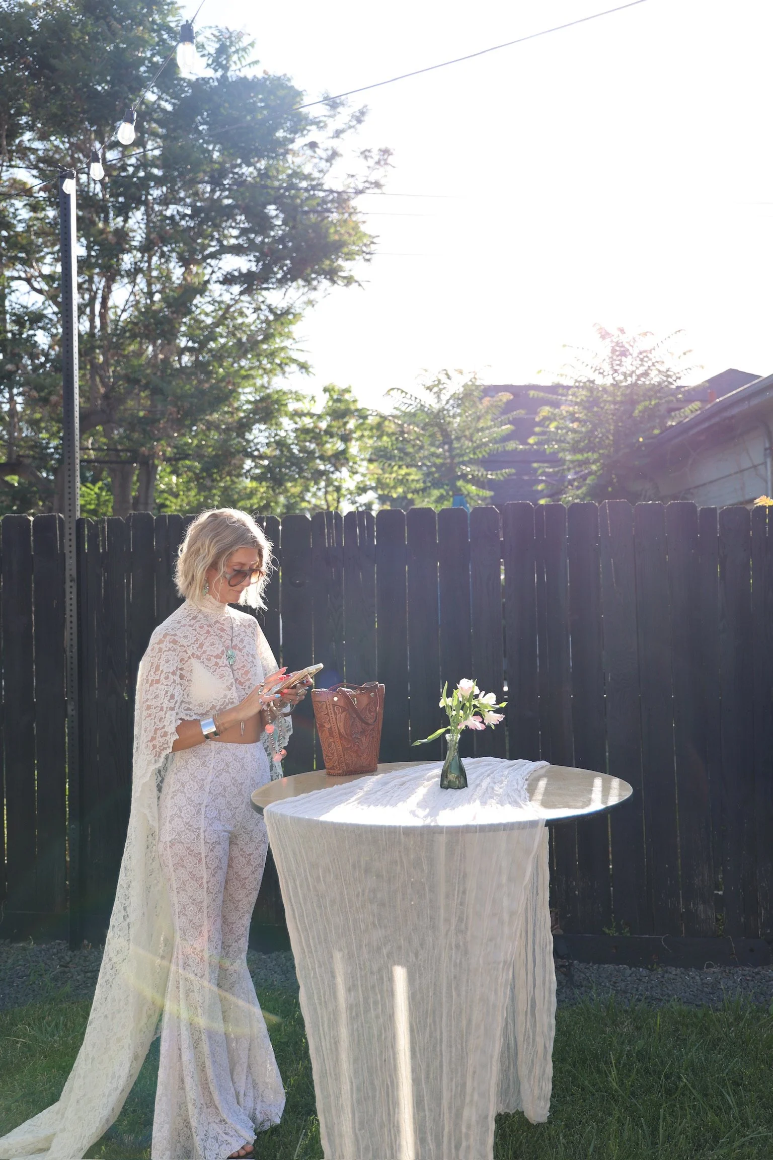 A woman in a white lace dress stands at an outdoor table with a vase of flowers, using a phone, in a backyard with a black wooden fence, trees, and string lights, under bright sunlight.