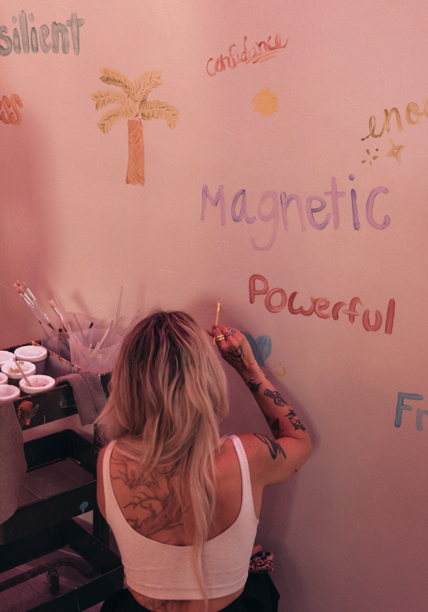 A woman with blonde hair and tattoos writing colorful words on a pink wall. The words include "Magnetic" and "Powerful". There are also drawings of a palm tree and small symbols, and containers of paint and brushes nearby.