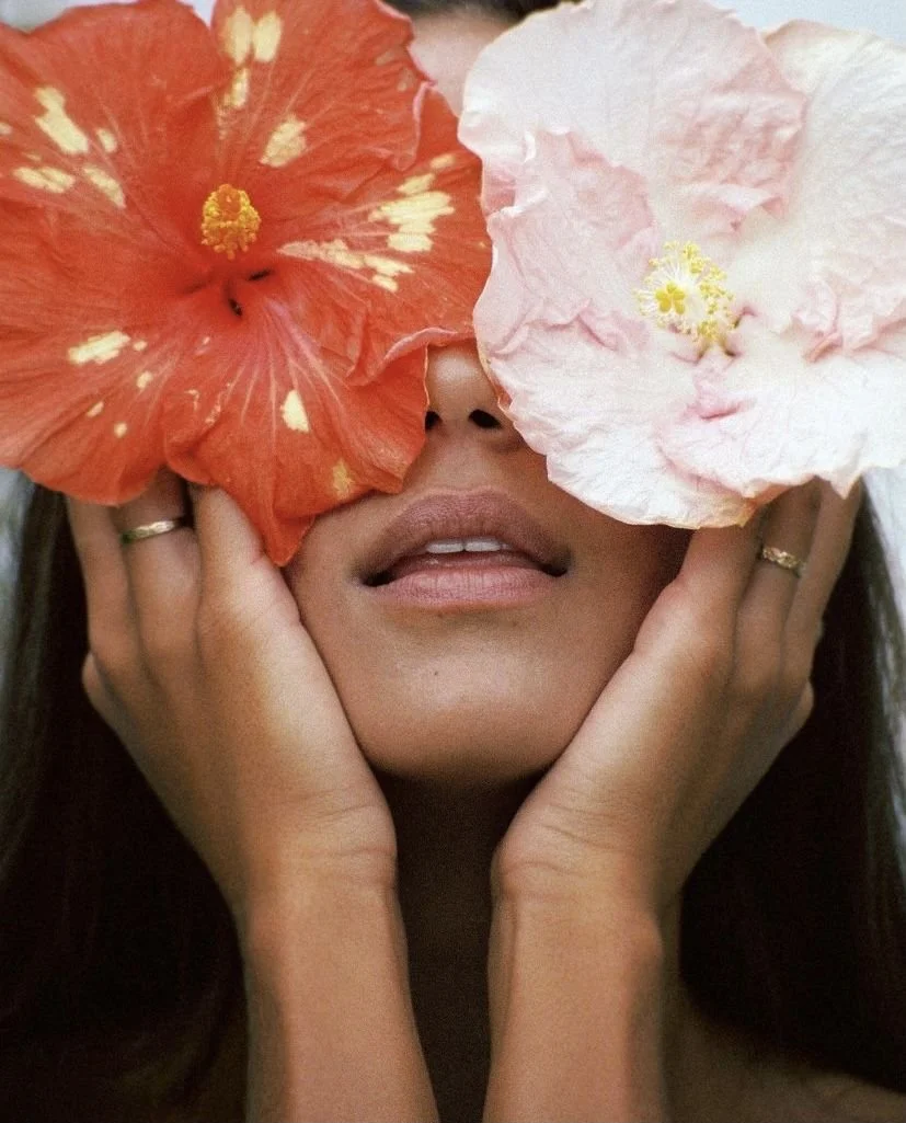 A woman holding two large flowers over her eyes, one orange with white spots and one pale pink, while her lips and hands are visible.