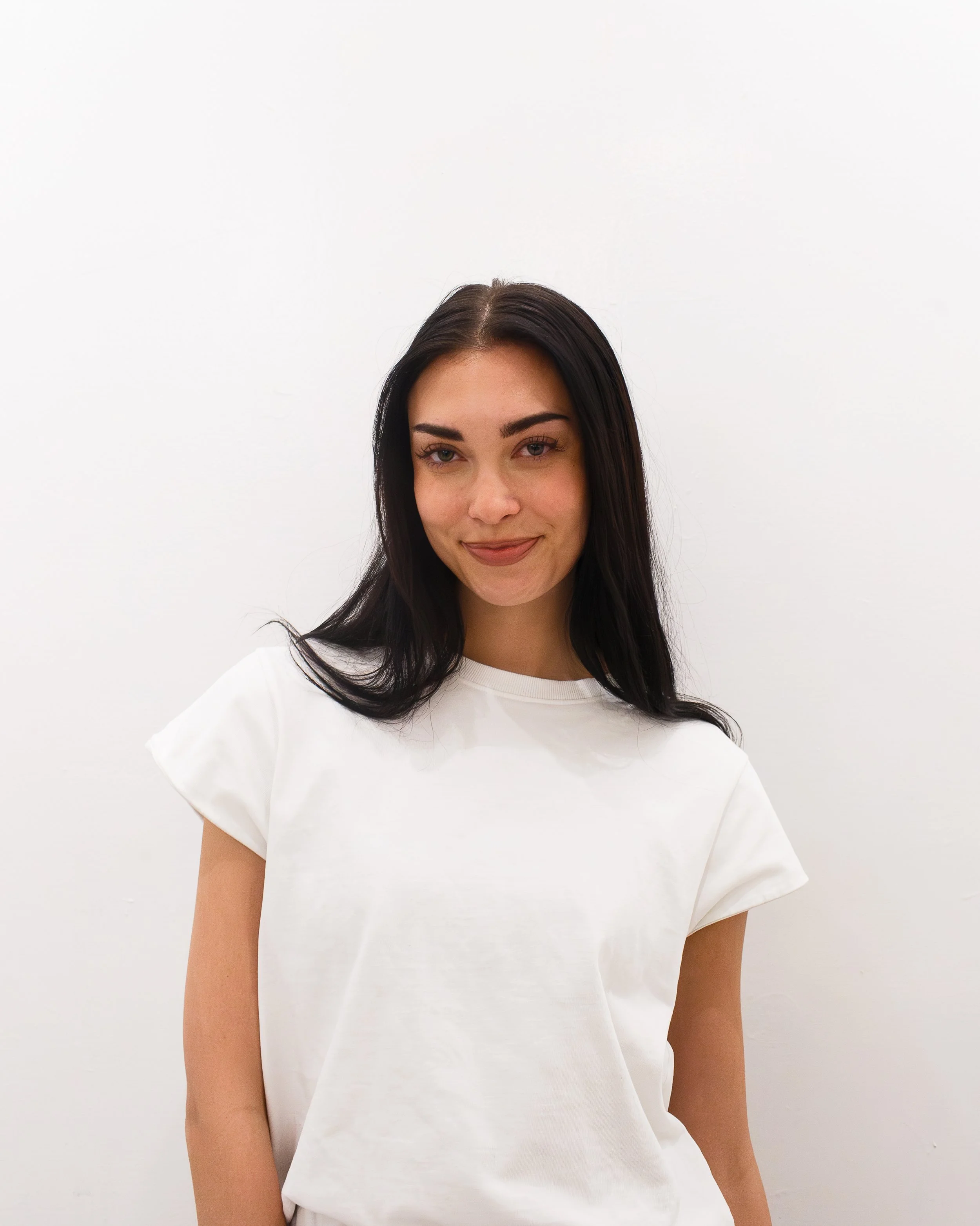 A young woman with long dark hair and a white t-shirt standing against a plain white background.