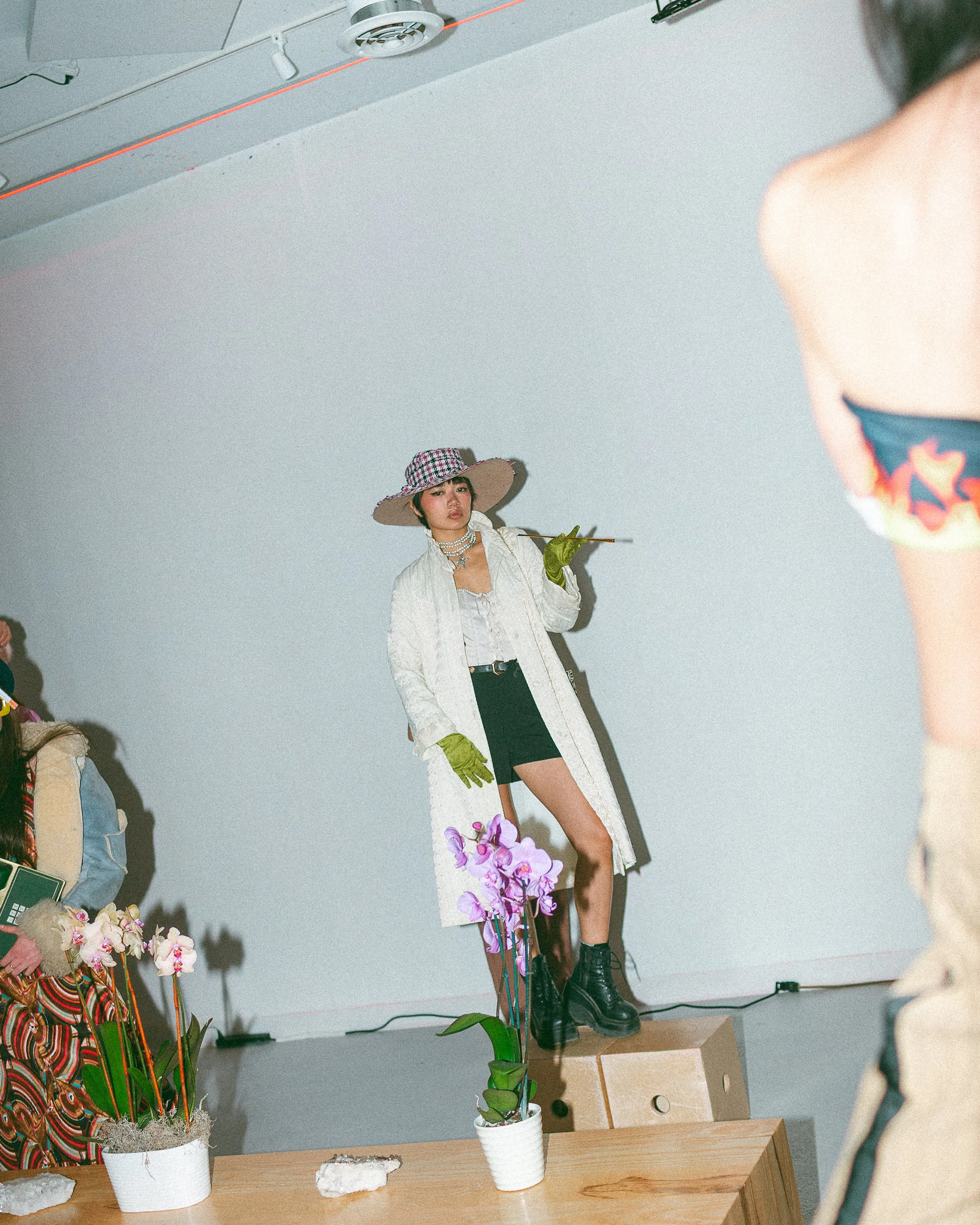 A woman is standing on a small stage, wearing a black hat with a colorful pattern, a white jacket, a black mini skirt, and black combat boots. She is holding a single pink orchid flower and has a neutral expression. There are potted orchids on a wooden table in the foreground and a mannequin dressed in colorful clothing on the left side. The background is a plain white wall with some visible ceiling fixtures.
