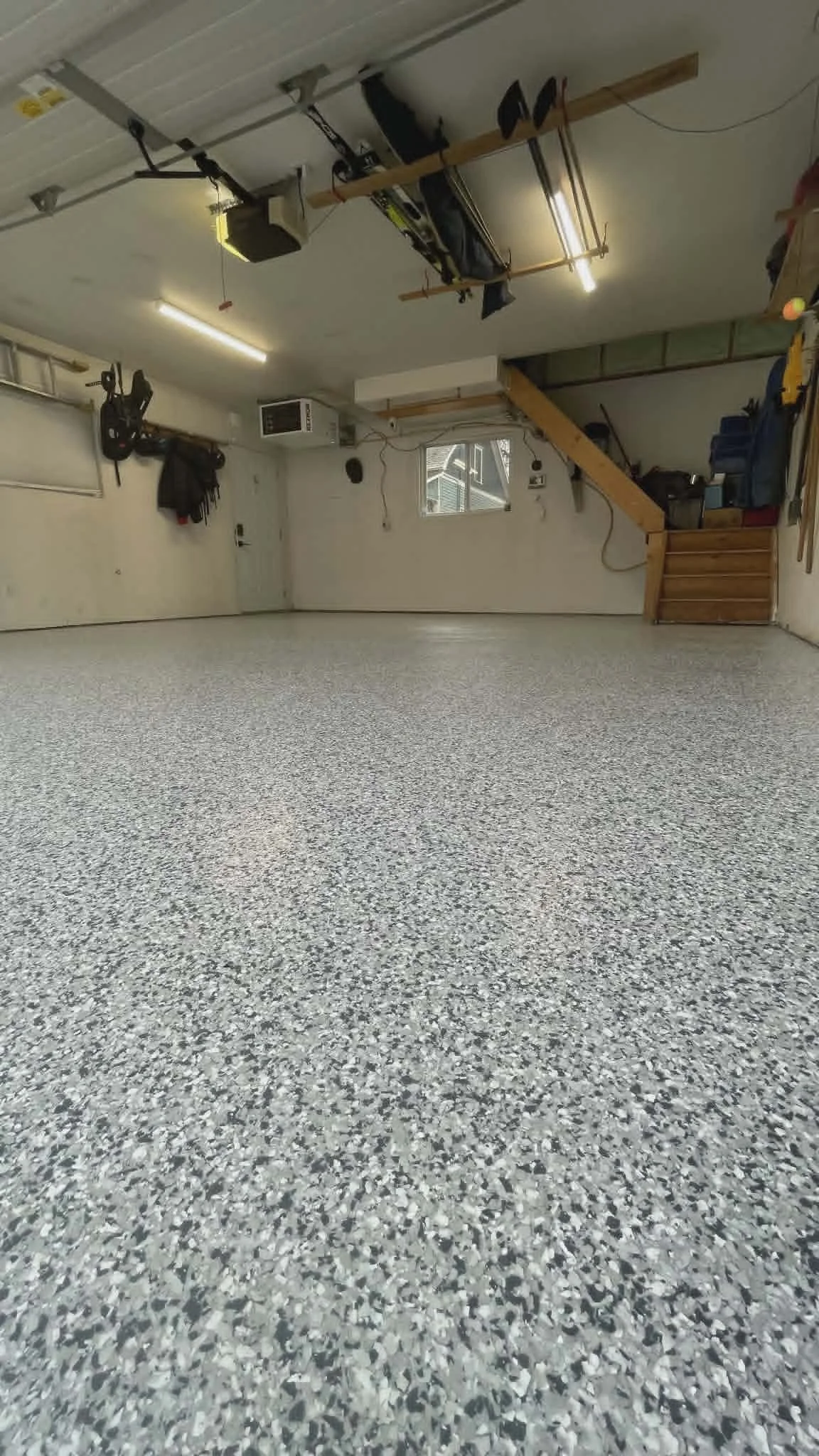 Empty garage with a speckled gray and white epoxy floor, overhead garage door, tools, and storage in the back.