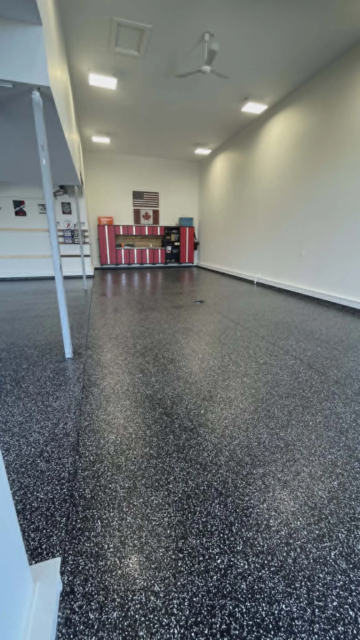 Empty room with speckled black and white flooring, white walls, a ceiling fan, ceiling lights, an American flag, a Canadian flag, and a red vending machine.