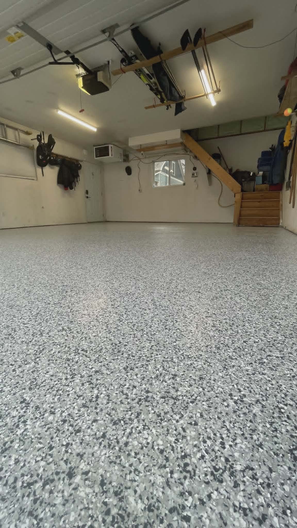 Empty garage with speckled gray floor, white walls, small window, and various tools and equipment hanging or stored.