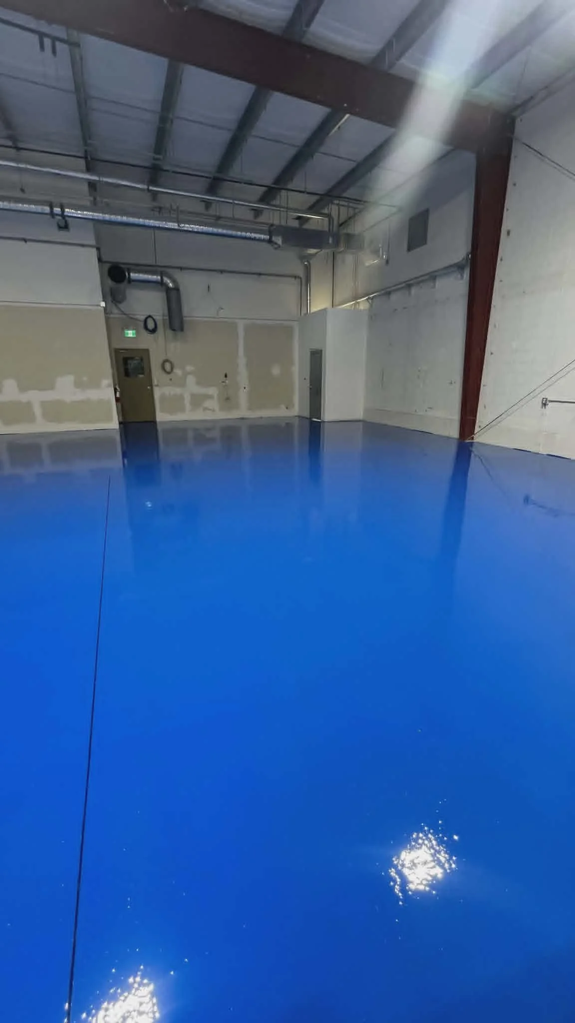 Empty indoor warehouse or factory with blue epoxy-coated floor, unfinished beige walls, exposed ductwork, and metal support beams.