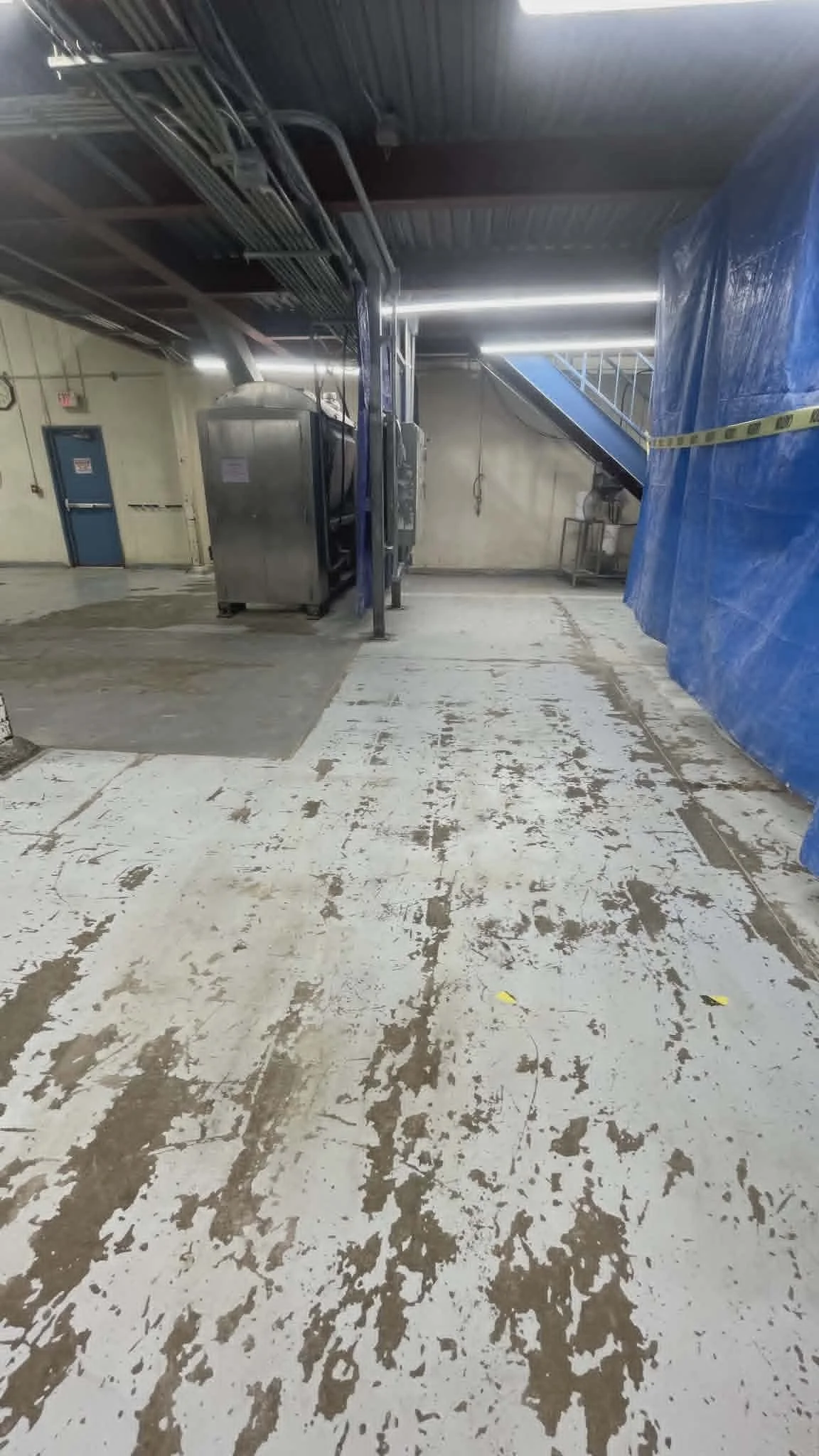 An industrial loading or storage area with worn-out white and brown painted concrete floor, metal equipment, blue tarps, and fluorescent lighting.