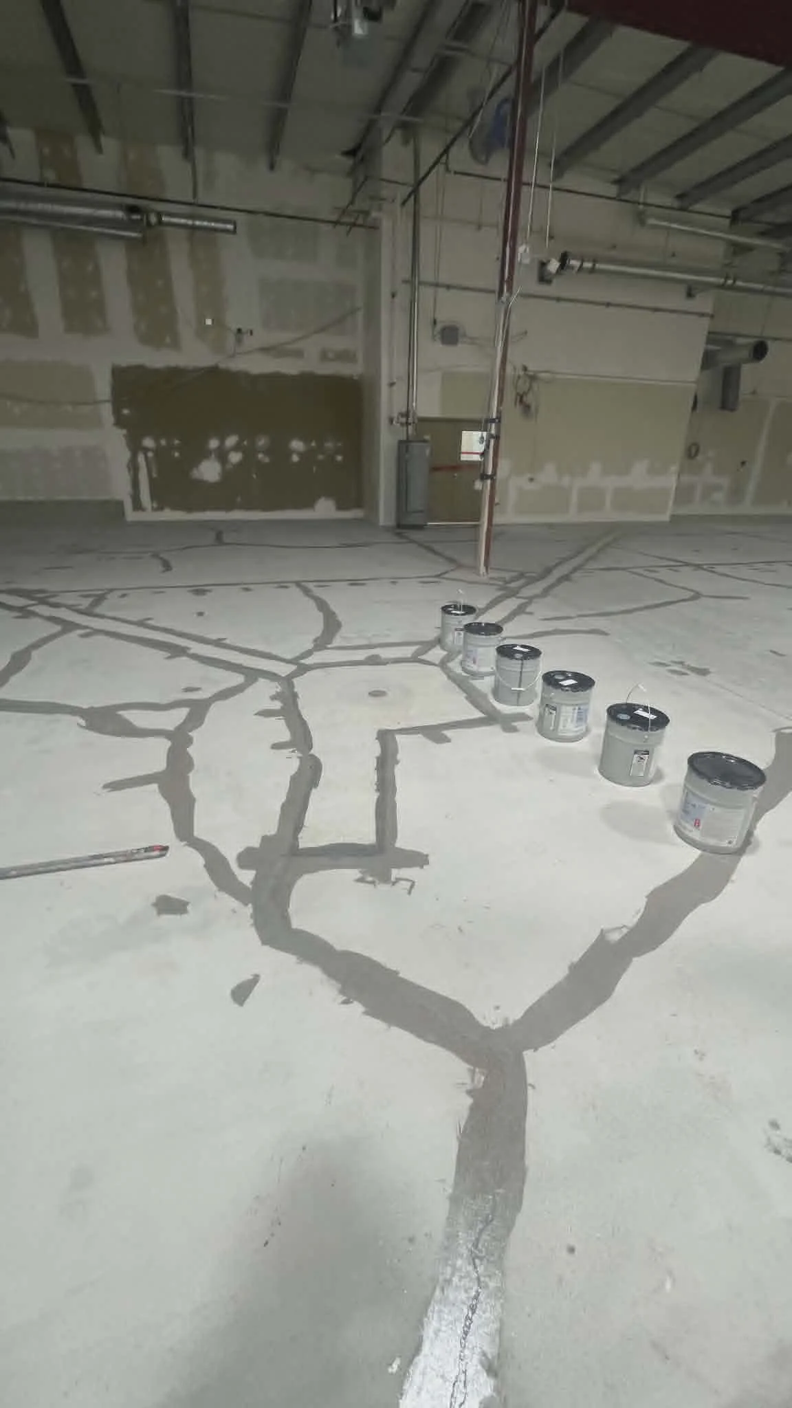 Construction site with cracked concrete floor and multiple cans of paint, unfinished walls, exposed pipes and ceiling.