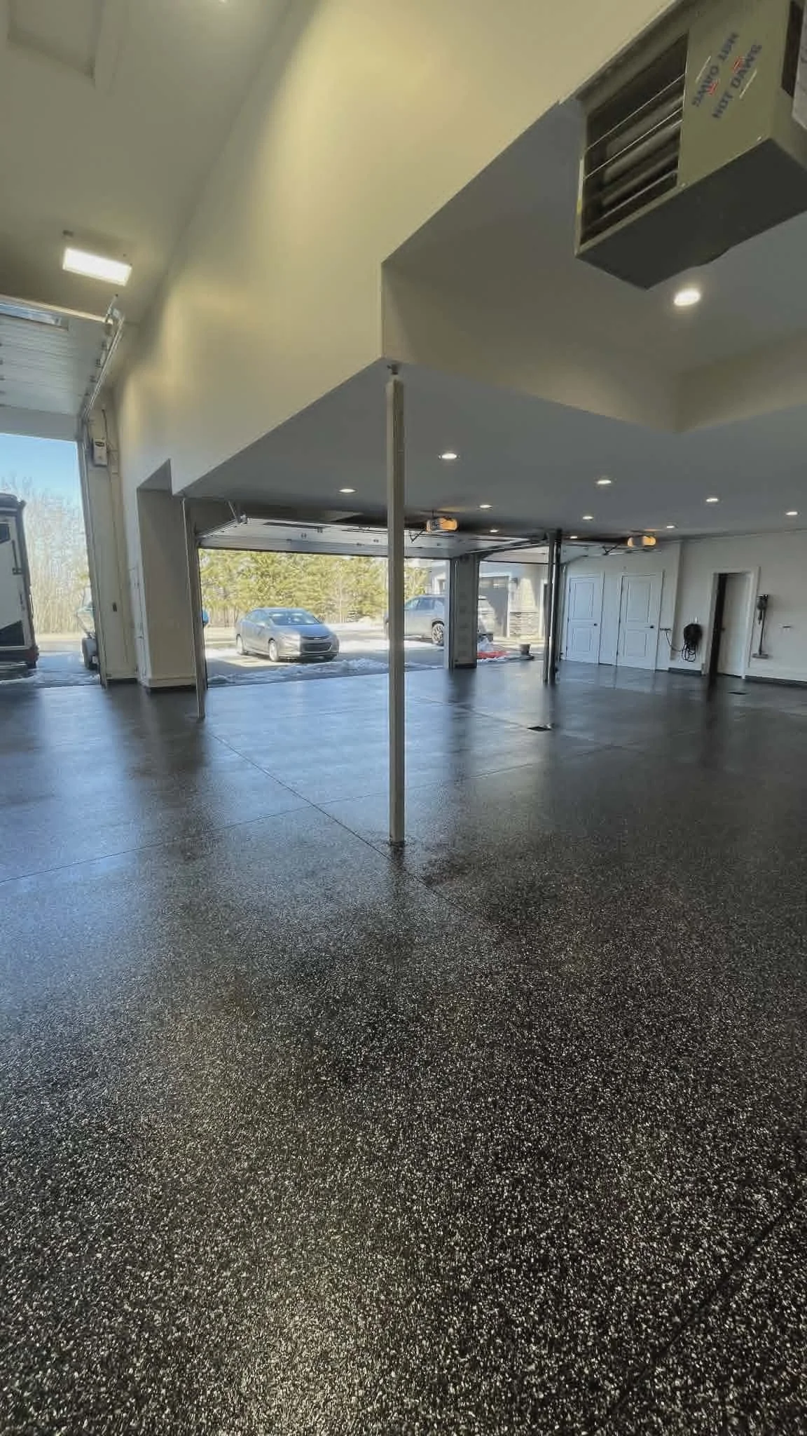 Empty garage with speckled epoxy floor, open garage doors, parked cars outside, and ceiling lights.