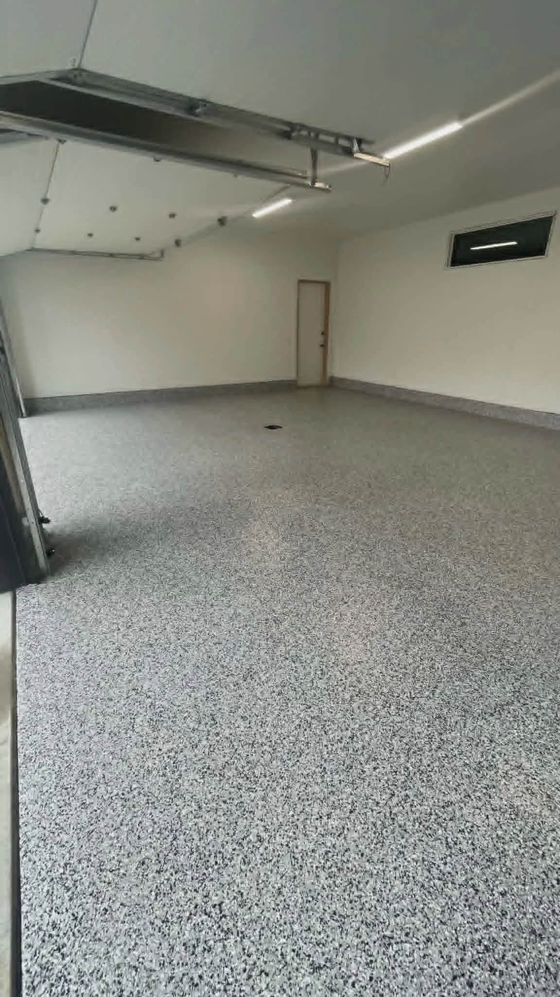 Empty garage with speckled gray flooring, a closed door, an air vent on the wall, and a partially open garage door with metal tracks.