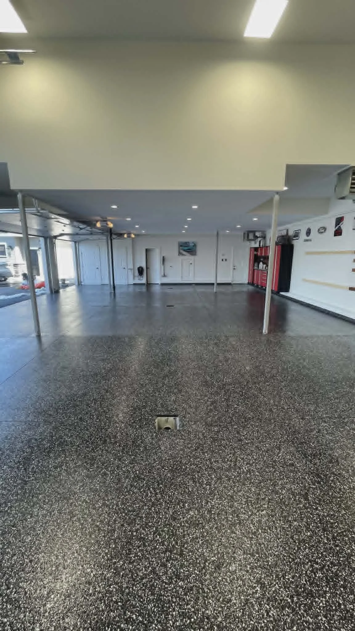 Empty room with a speckled black and white floor, white walls, and bright overhead lighting. There are some red tools or equipment on the right side and some windows and doors on the left.