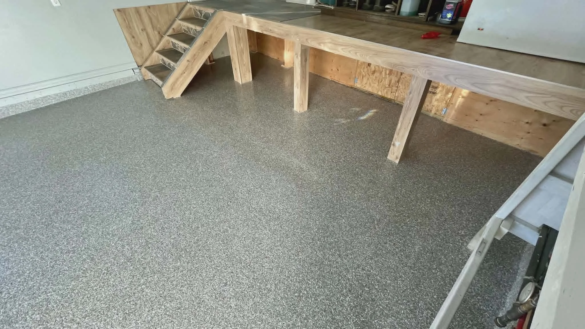 A corner of a room with a wooden countertop and a small wooden staircase with open risers leading to an elevated surface. The floor has speckled gray carpet. There are shelves and cabinets in the background, with various objects on top.