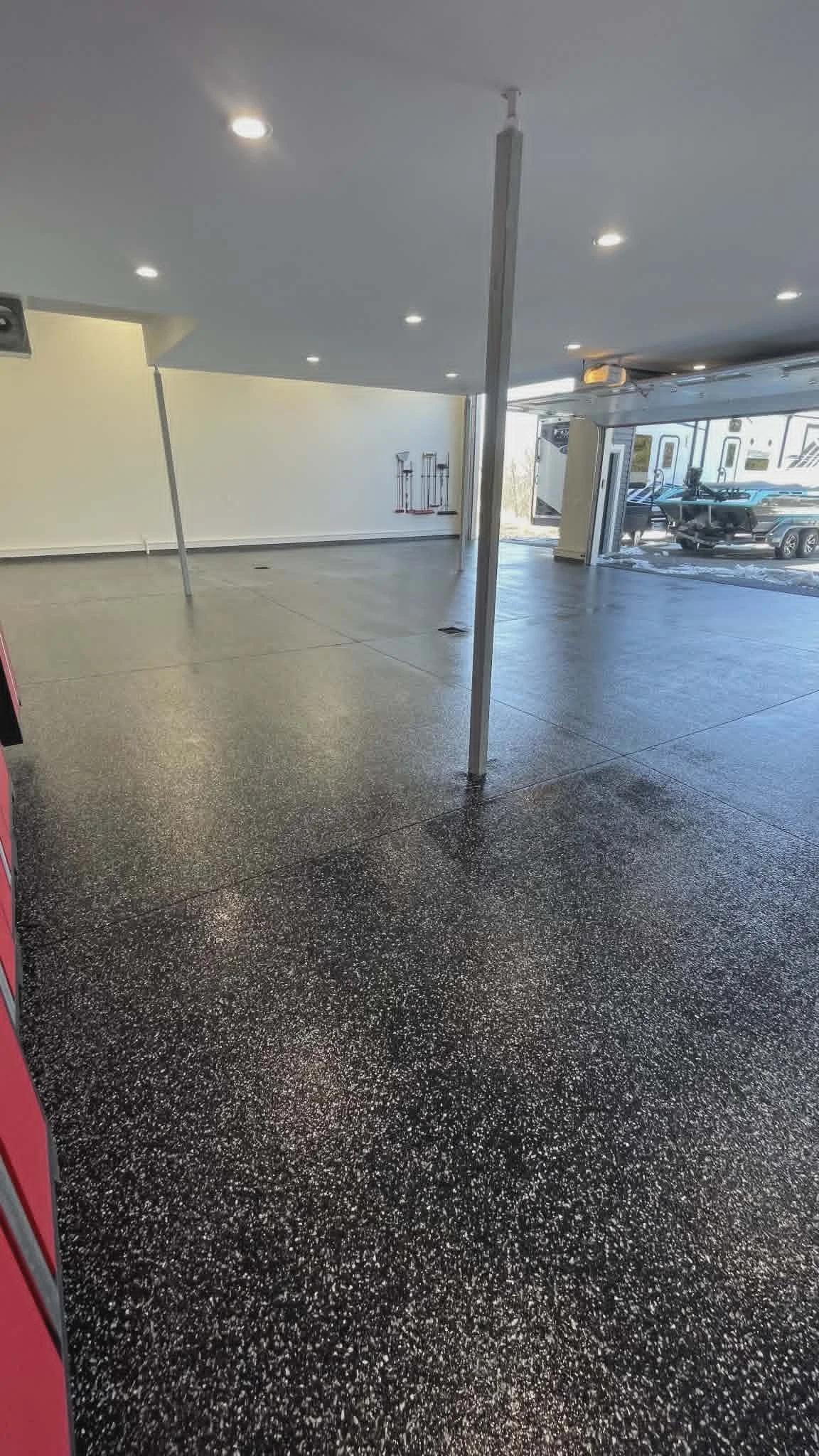 Empty indoor space with black and white speckled flooring, white walls, and ceiling-mounted lights. There are metal poles supporting the ceiling and a view of the outdoors through an open garage door, where vehicles and RVs are parked.
