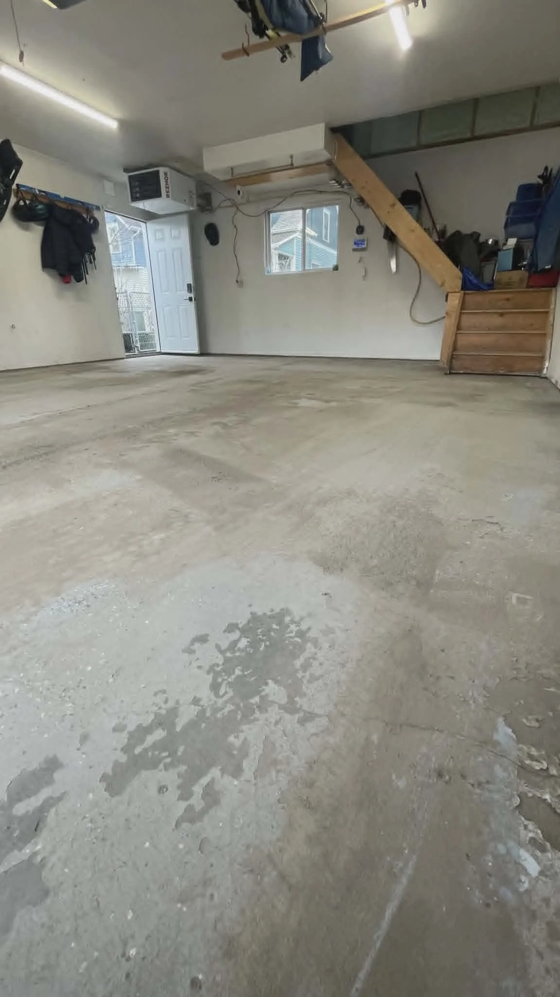Empty garage with a concrete floor and walls, door, window, and shelves with tools and items stored, with some hanging on the walls.