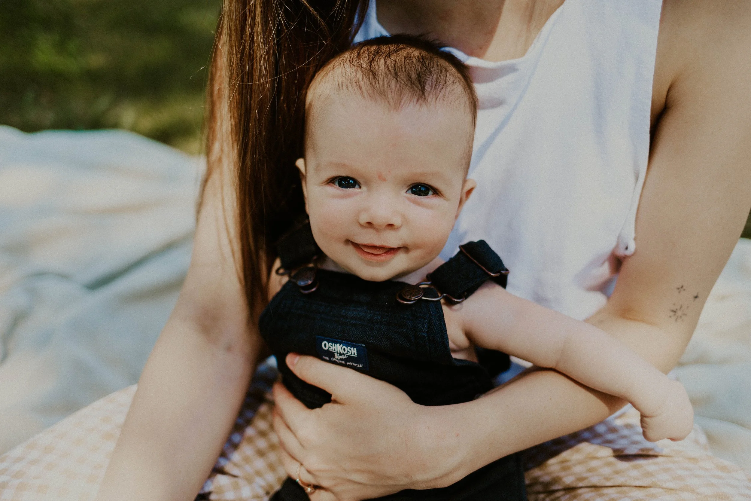 A young child with blue eyes and light brown hair being held by an adult, possibly a mother, with visible arms and a tattoo. The child is wearing a black OshKosh Baby Gap outfit with adjustable straps and a small logo. The background appears to be ou