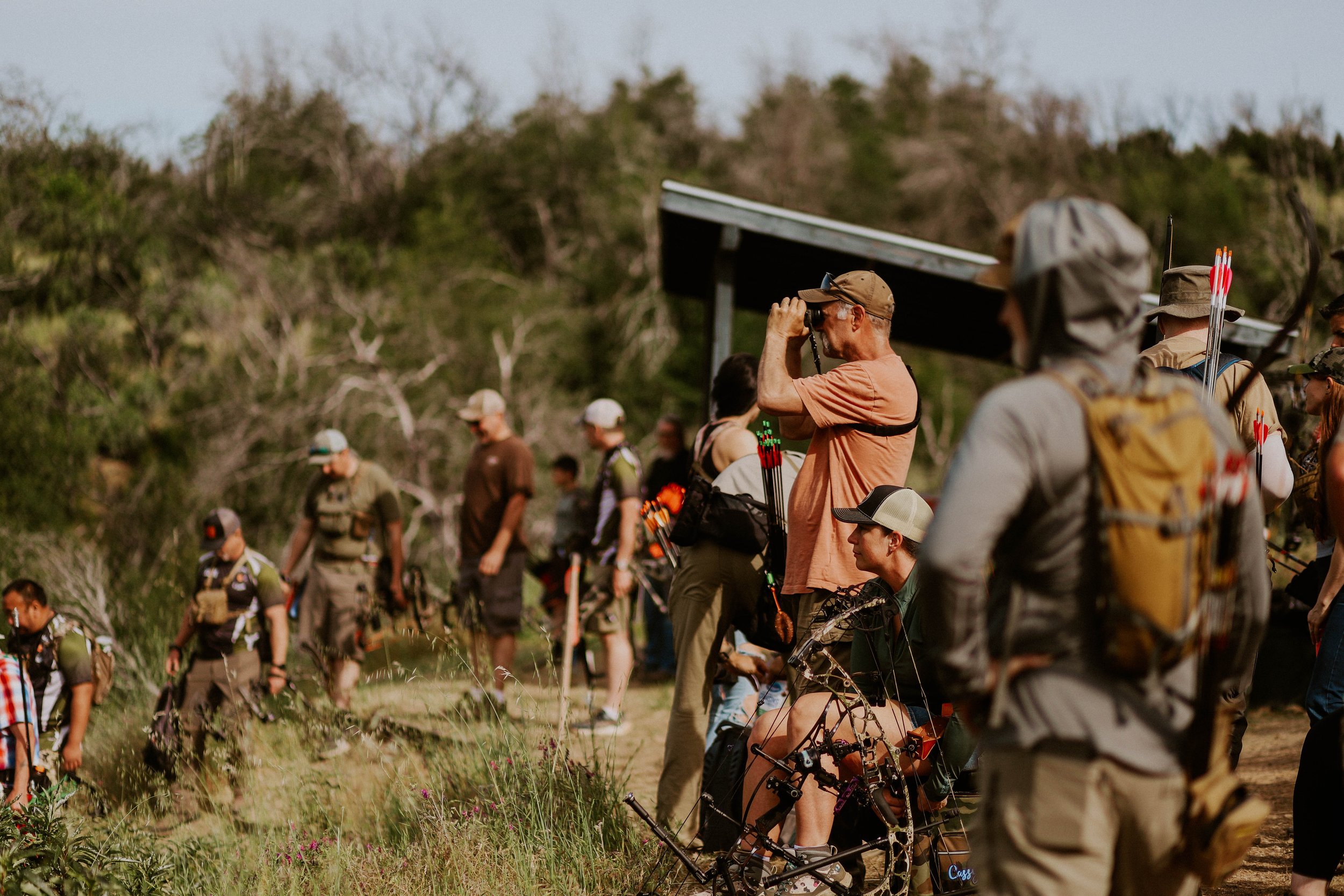 People in outdoor clothing, some with archery gear and arrows, gathered in a natural setting with trees, likely participating in a recreational or competitive archery event.